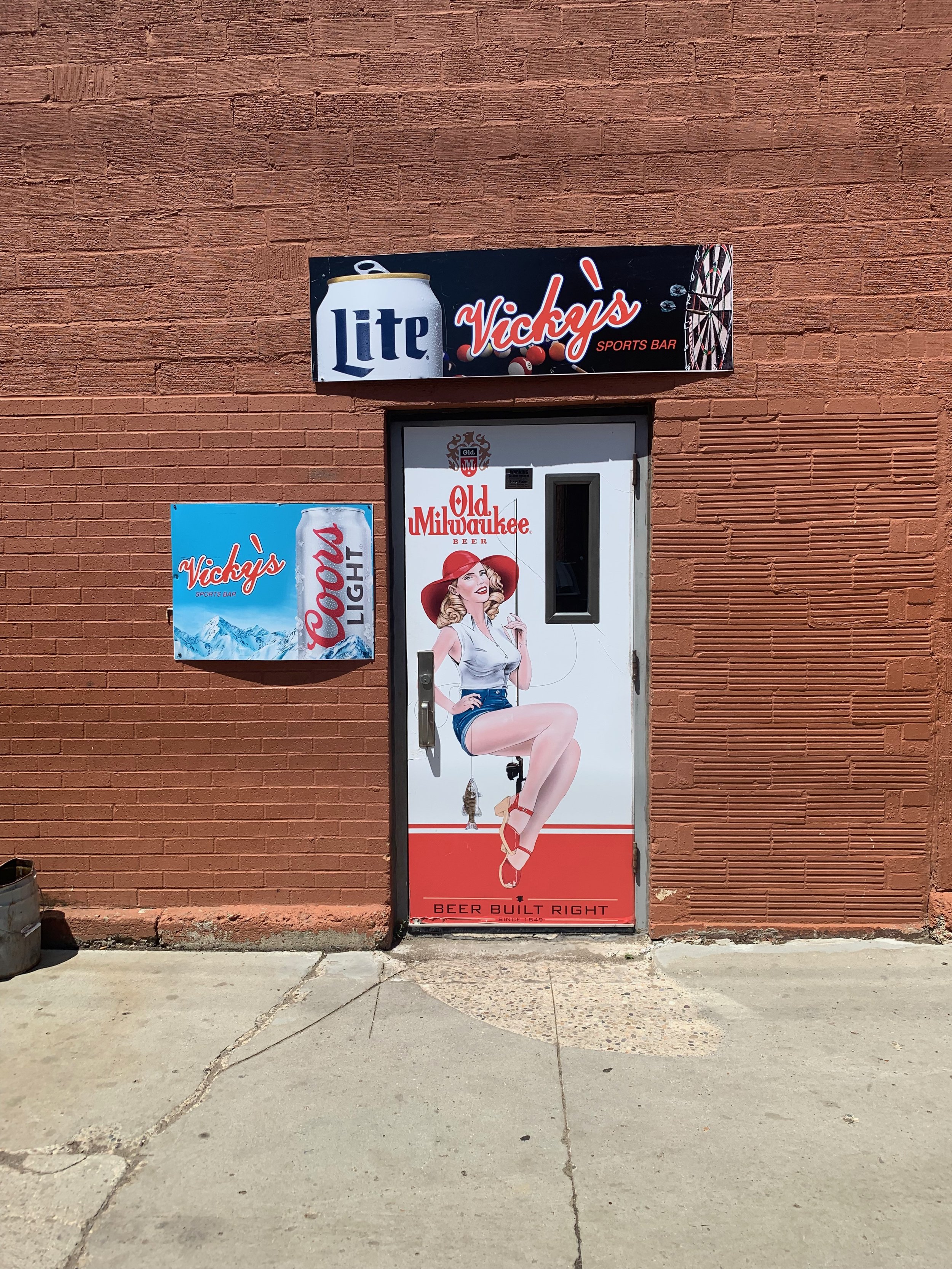 Door with a pin-up style woman holding a fishing pole, on a mural for Old Milwaukee beer. Signs for Vicky's sports bar, Miller Lite, and Coors Light are on the wall.