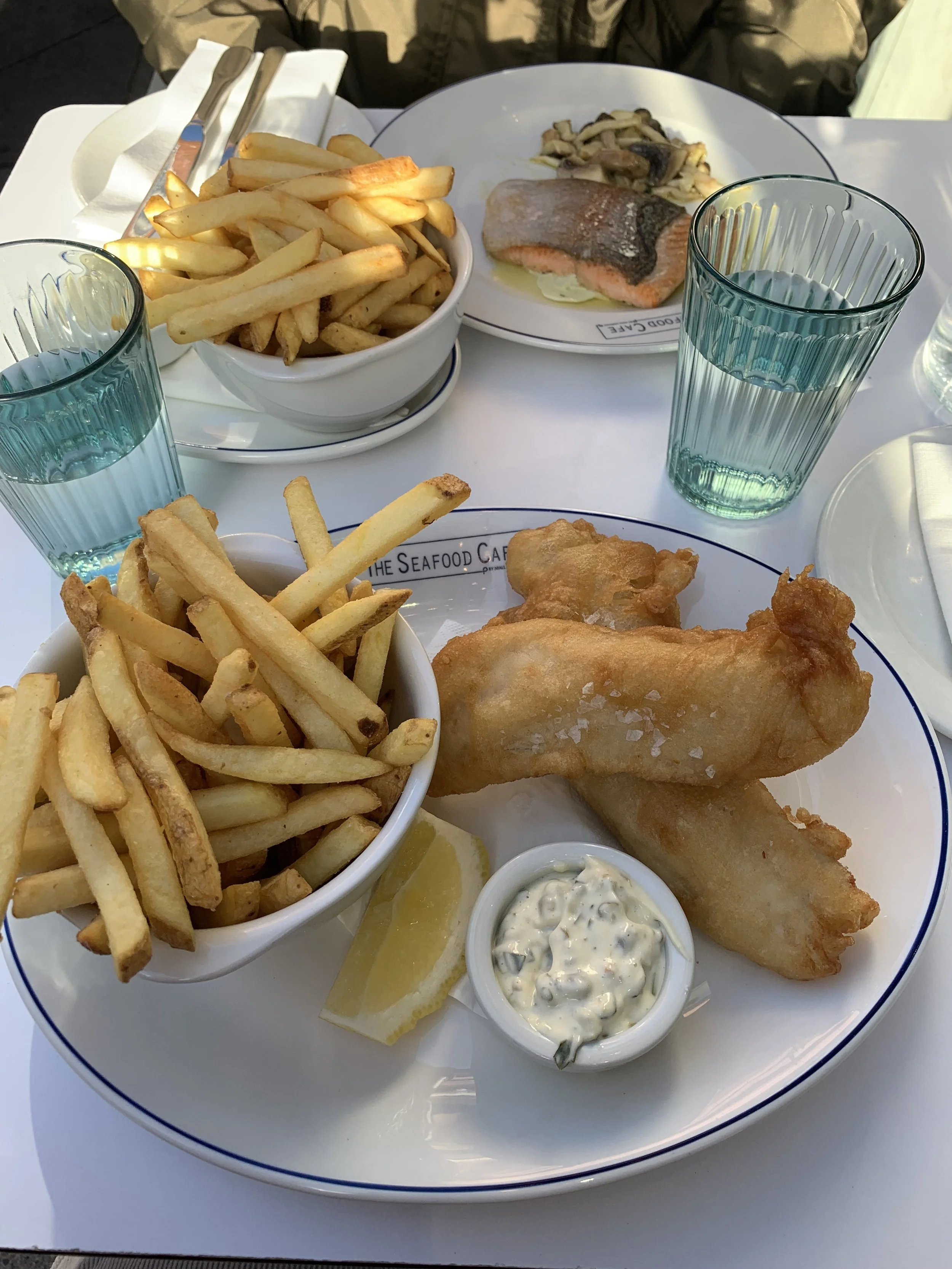 Fried fish and chips with tartar sauce, lemon wedge, and two bowls of French fries on a white table at a seafood restaurant.