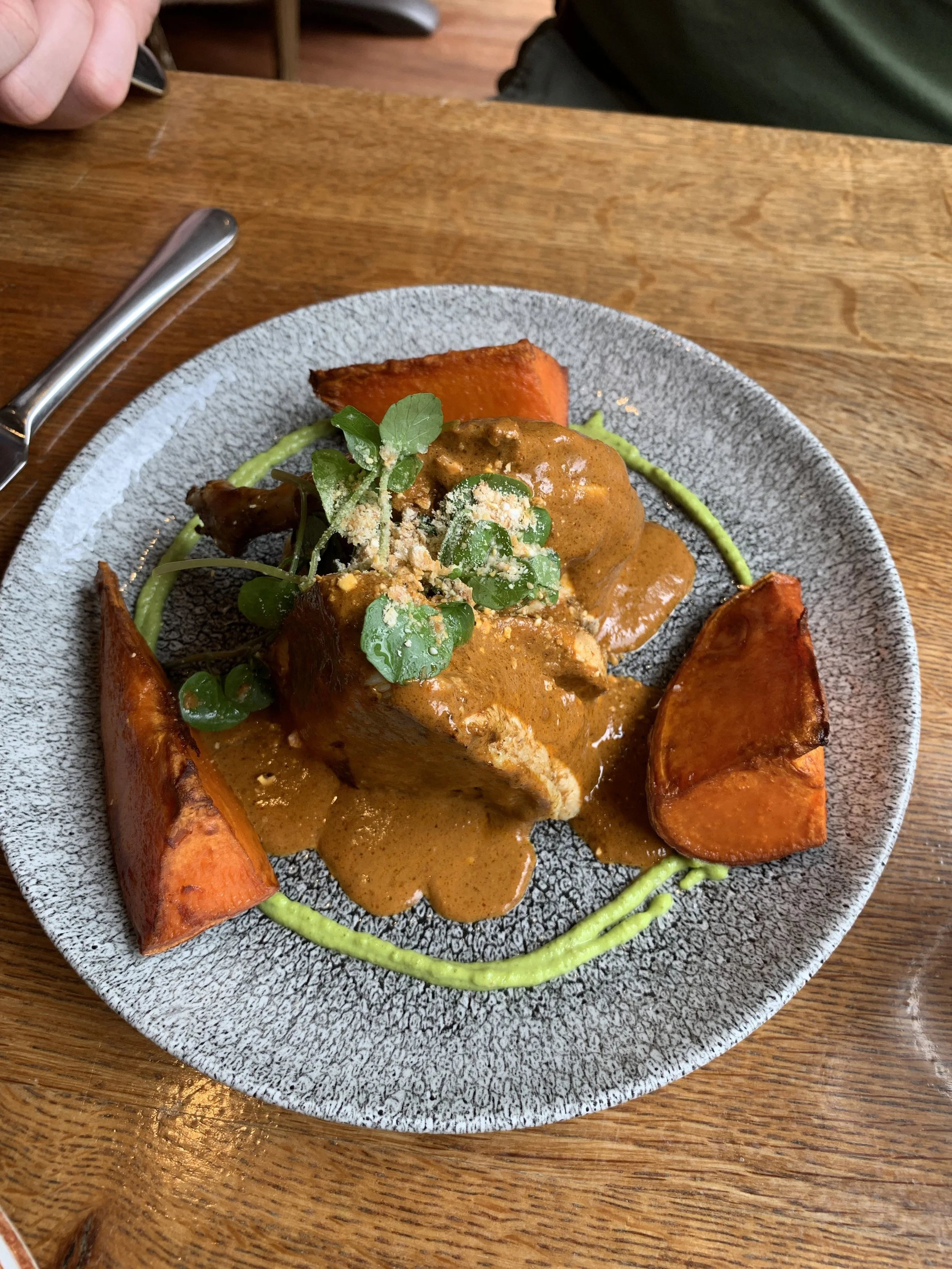 A plate of food featuring a piece of meat topped with sauce and garnished with herbs, served with roasted sweet potatoes and a ring of green puree on a speckled gray plate.