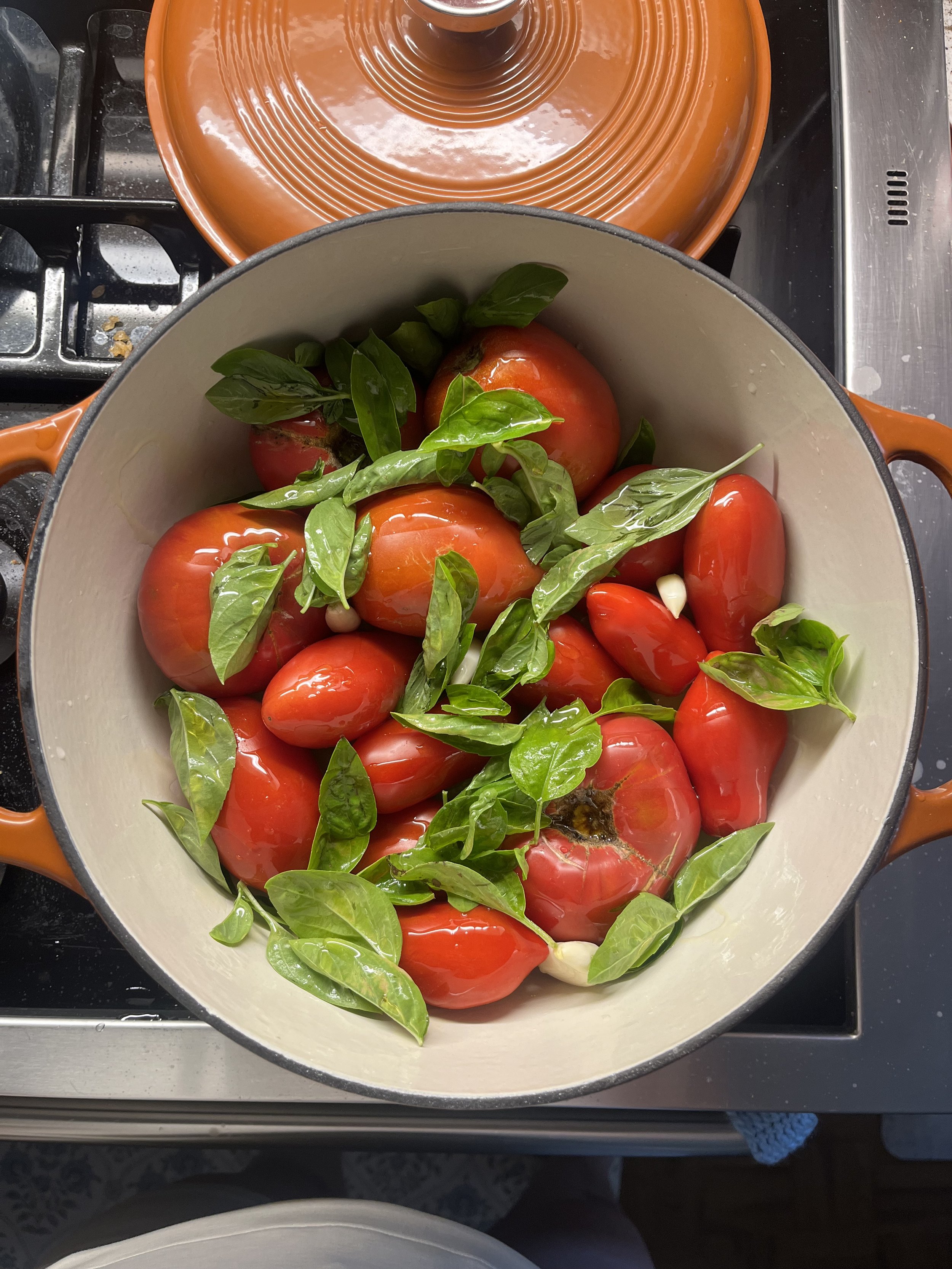 A Dutch oven filled with large red heirloom tomatoes, fresh basil leaves, and garlic cloves.