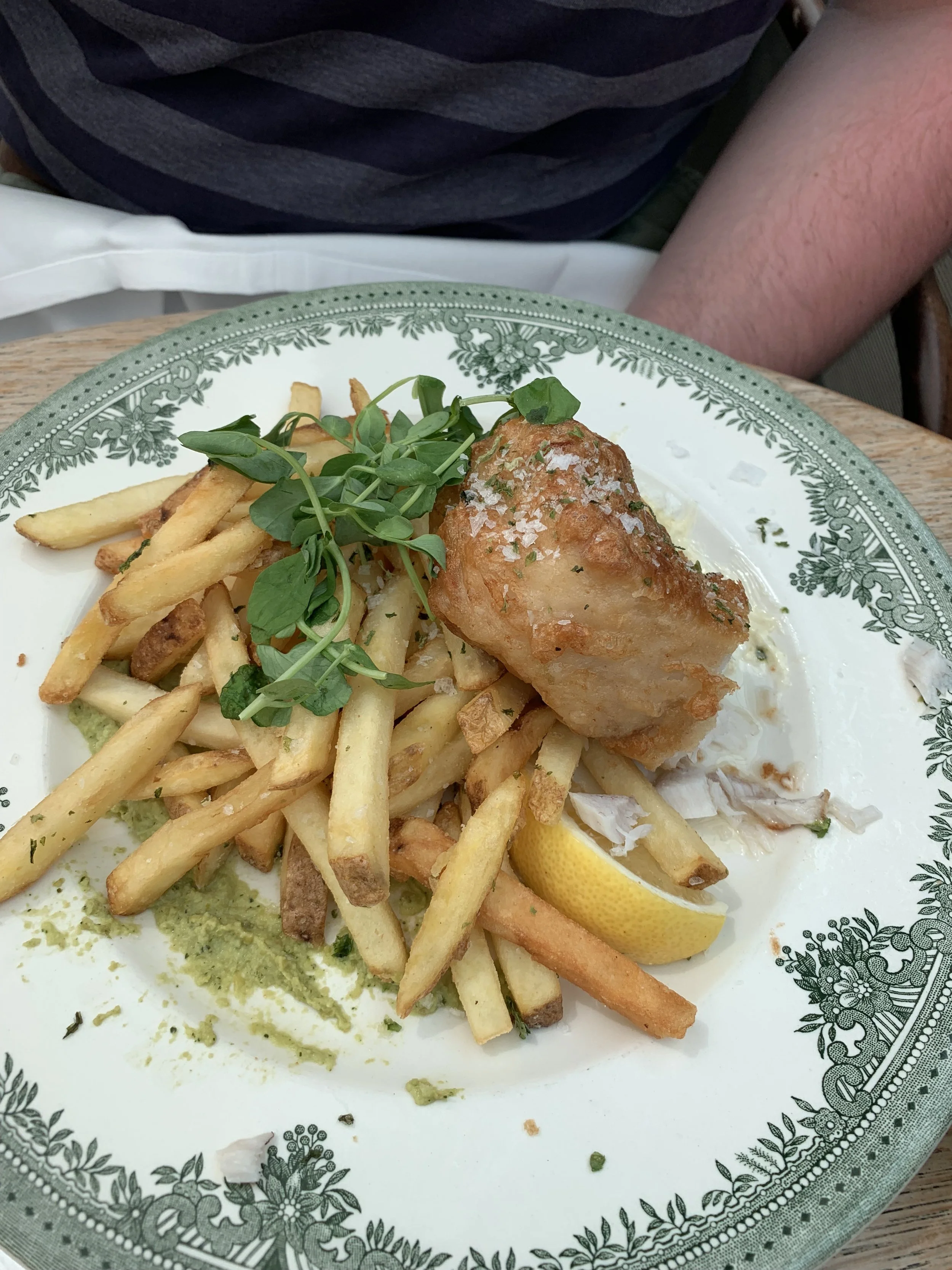 Plate of fish with French fries, lemon wedge, green sauce, and fresh herbs on a decorative plate.