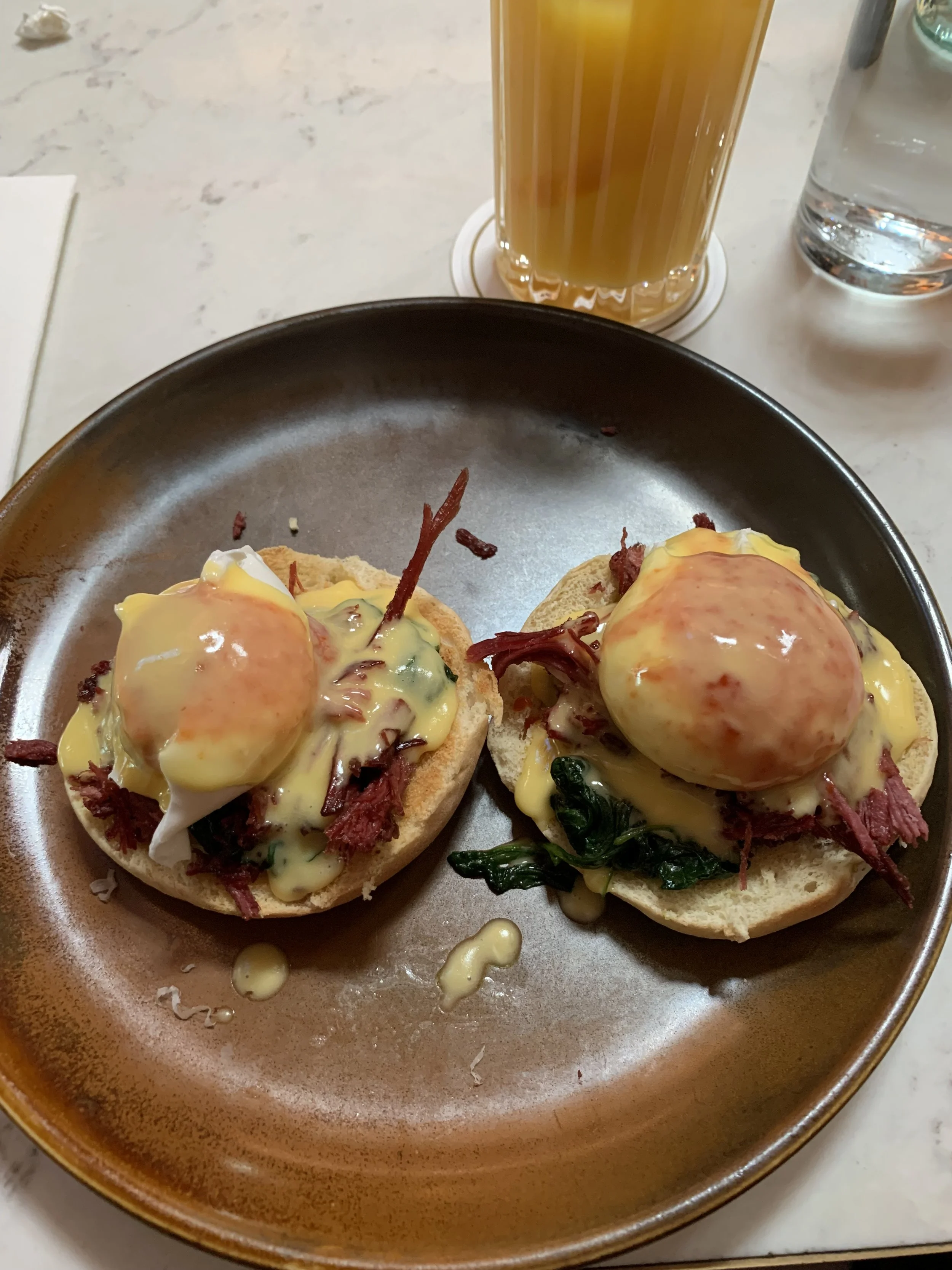 Two eggs Benedict with smoked salmon on English muffins, topped with hollandaise sauce, served on a brown plate. In the background, there is a glass of iced tea and a glass of water.