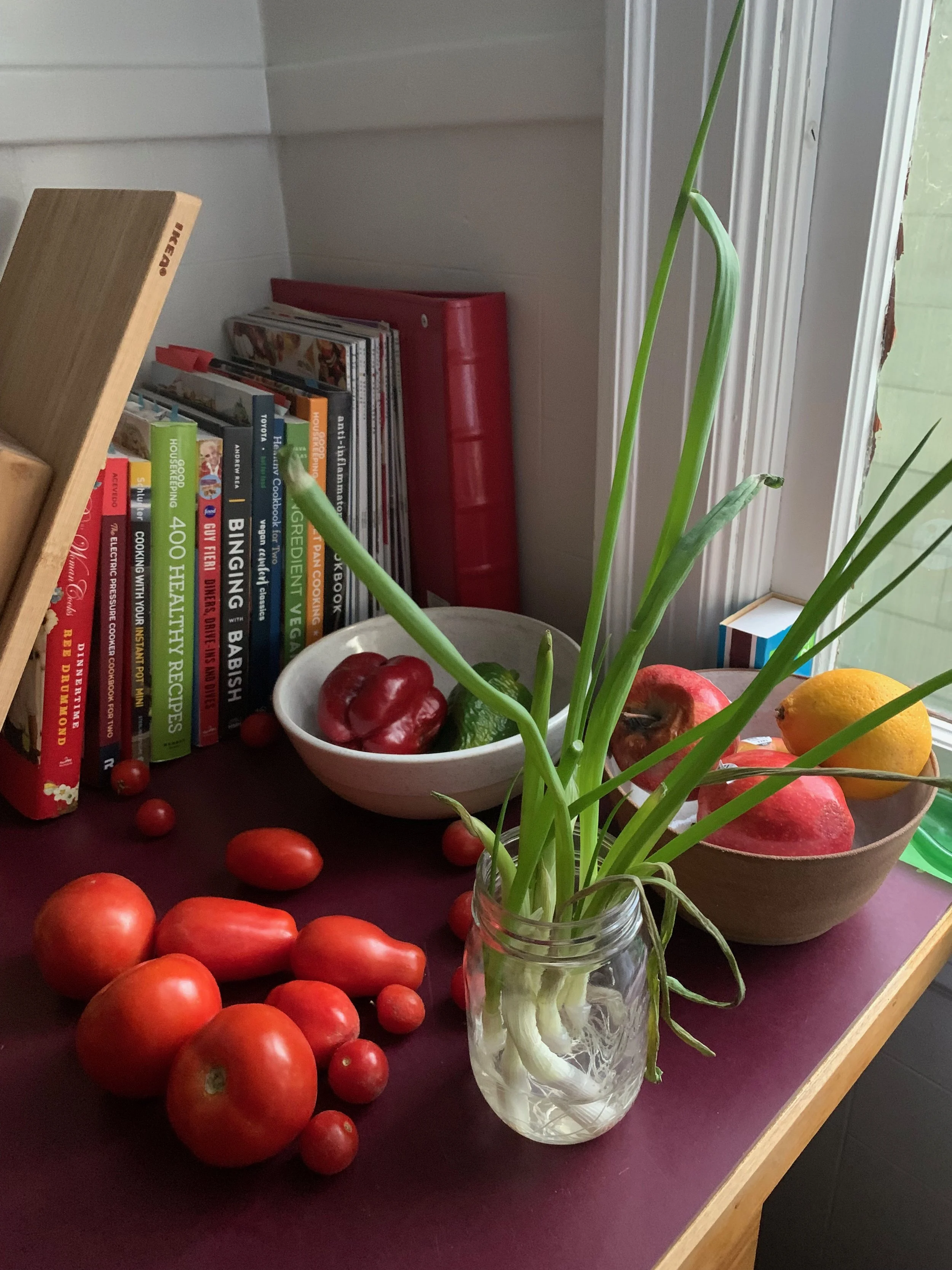A windowsill with a jar of green onion sprigs, bowls of tomatoes, peppers, cucumbers, an apple, lemon, and pomegranate, and a collection of cookbooks and magazines.