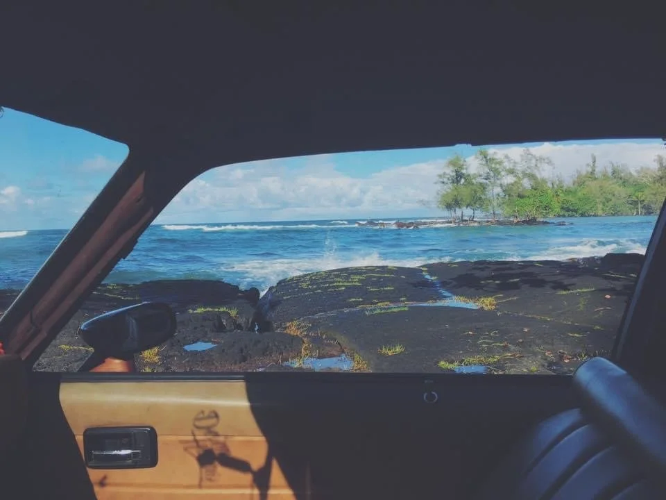 View from inside a vehicle showing a rocky shoreline with the ocean and small trees in the background on a clear day.