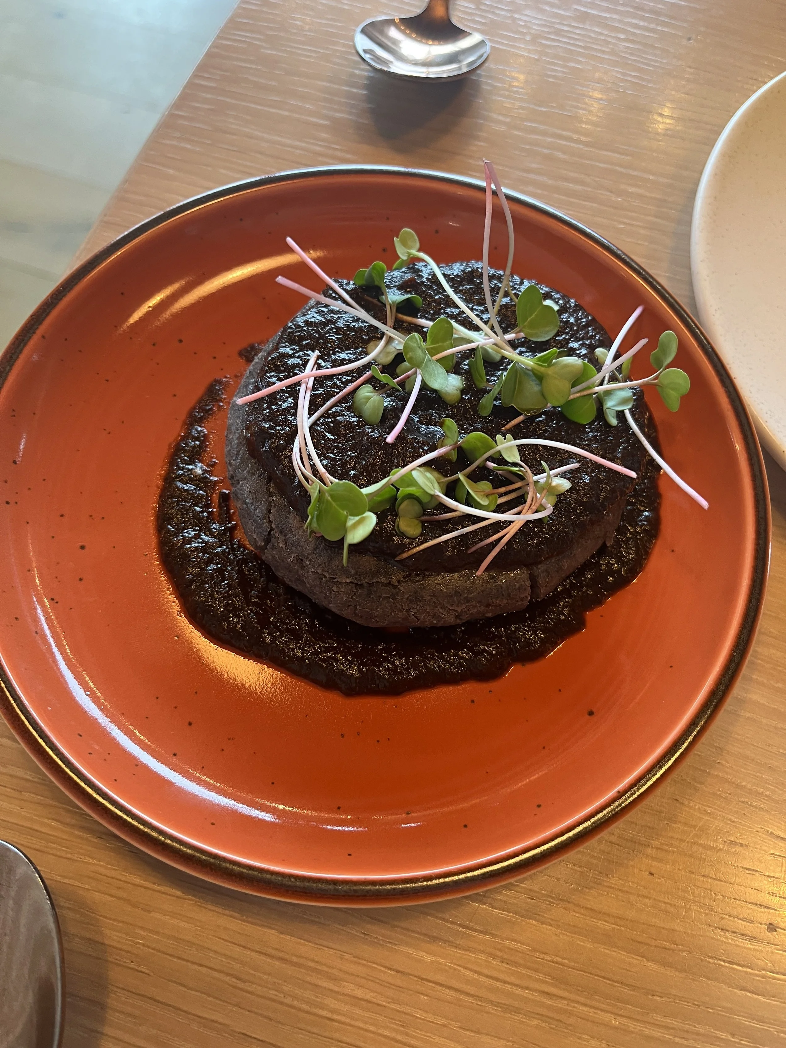 A serving of black food, possibly a black burger or sandwich, topped with microgreens, on an orange plate with black specks, on a wooden table.