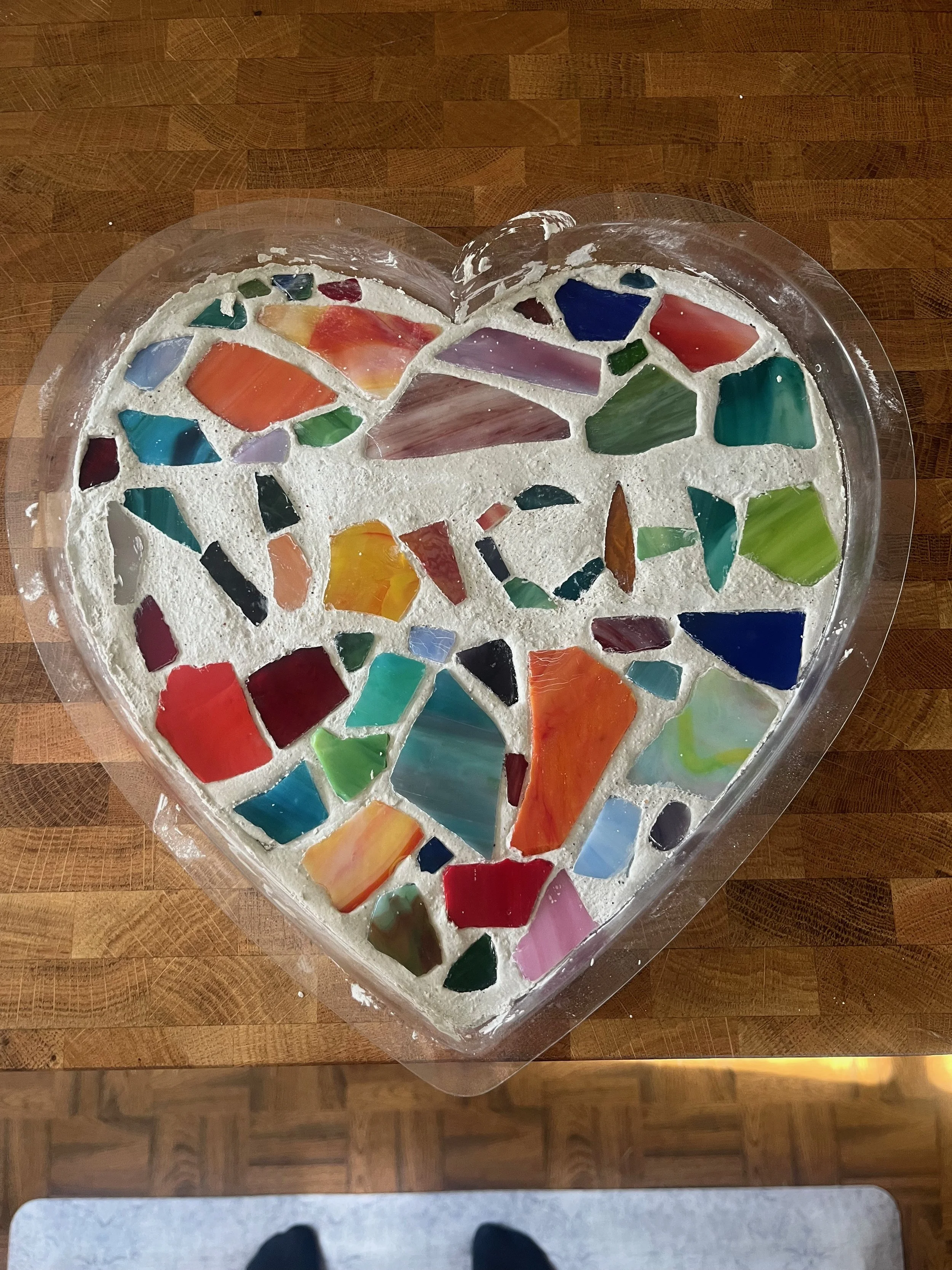 Heart-shaped mosaic art piece made with colorful stained glass pieces on a white base inside a clear plastic frame, placed on a wooden surface.