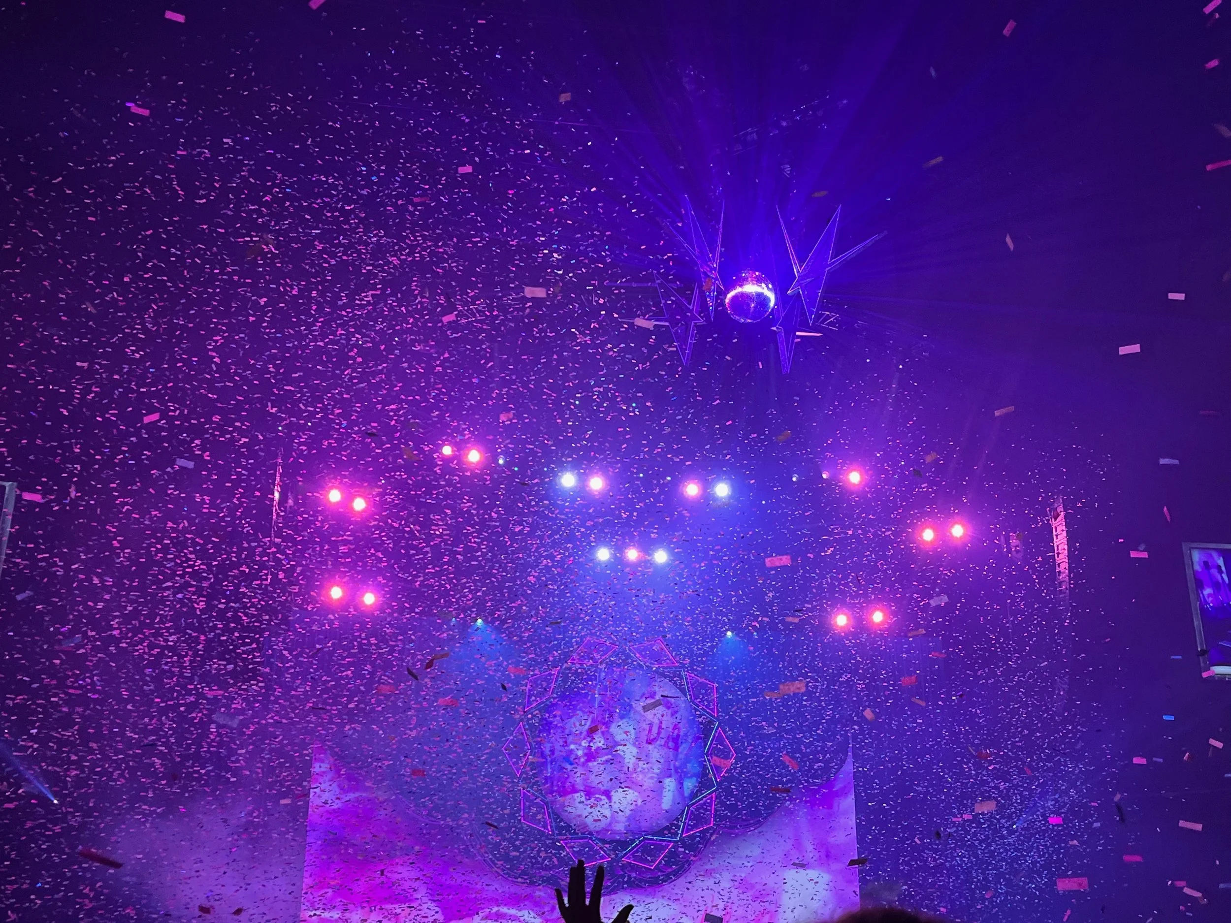 A vibrant concert scene with purple and pink lighting, confetti falling, and a DJ or performer illuminated on stage under a large disco ball.