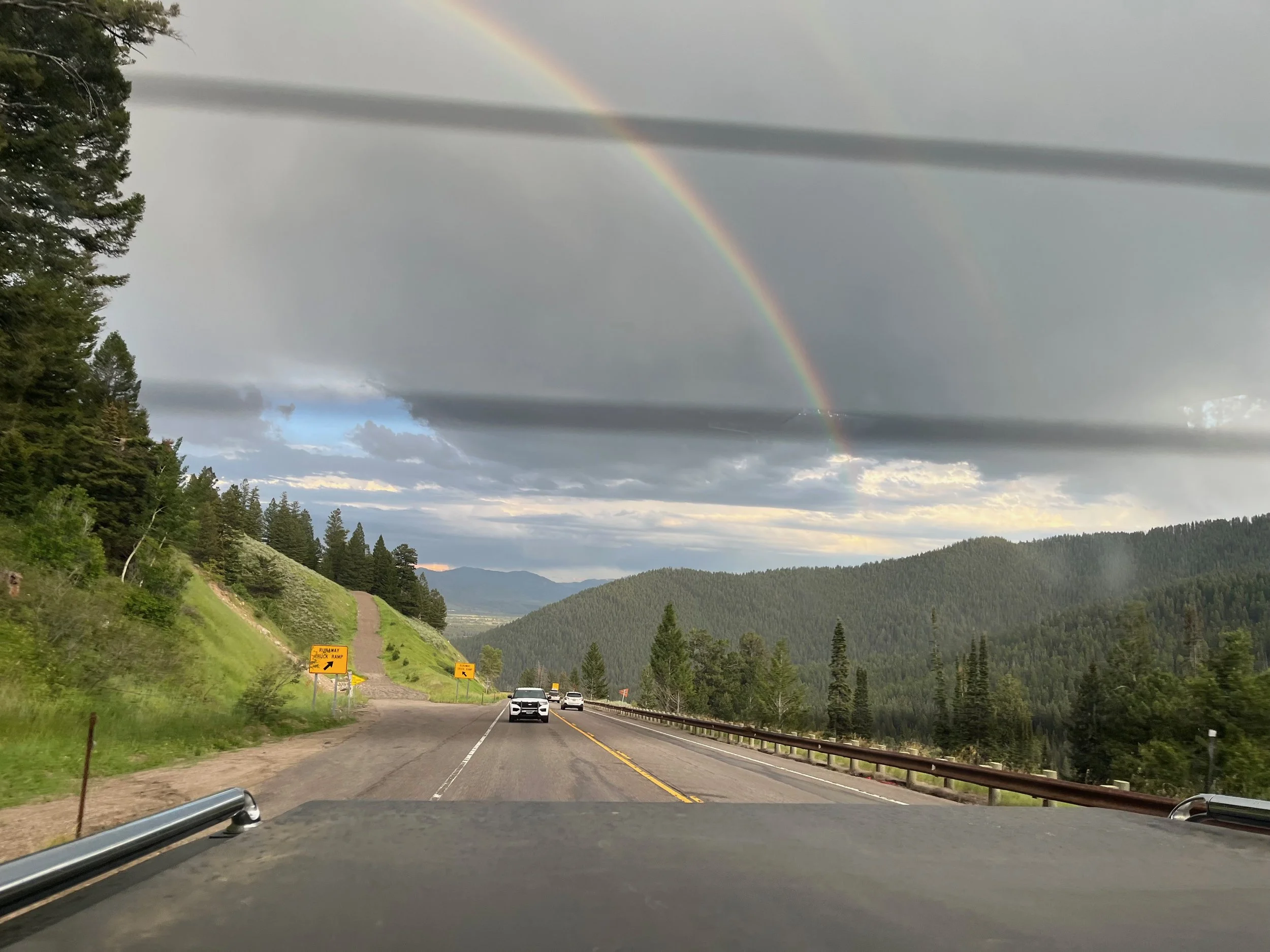 A scenic mountain landscape with a rainbow in the cloudy sky, viewed from a moving vehicle on a two-lane road surrounded by green trees and hills.