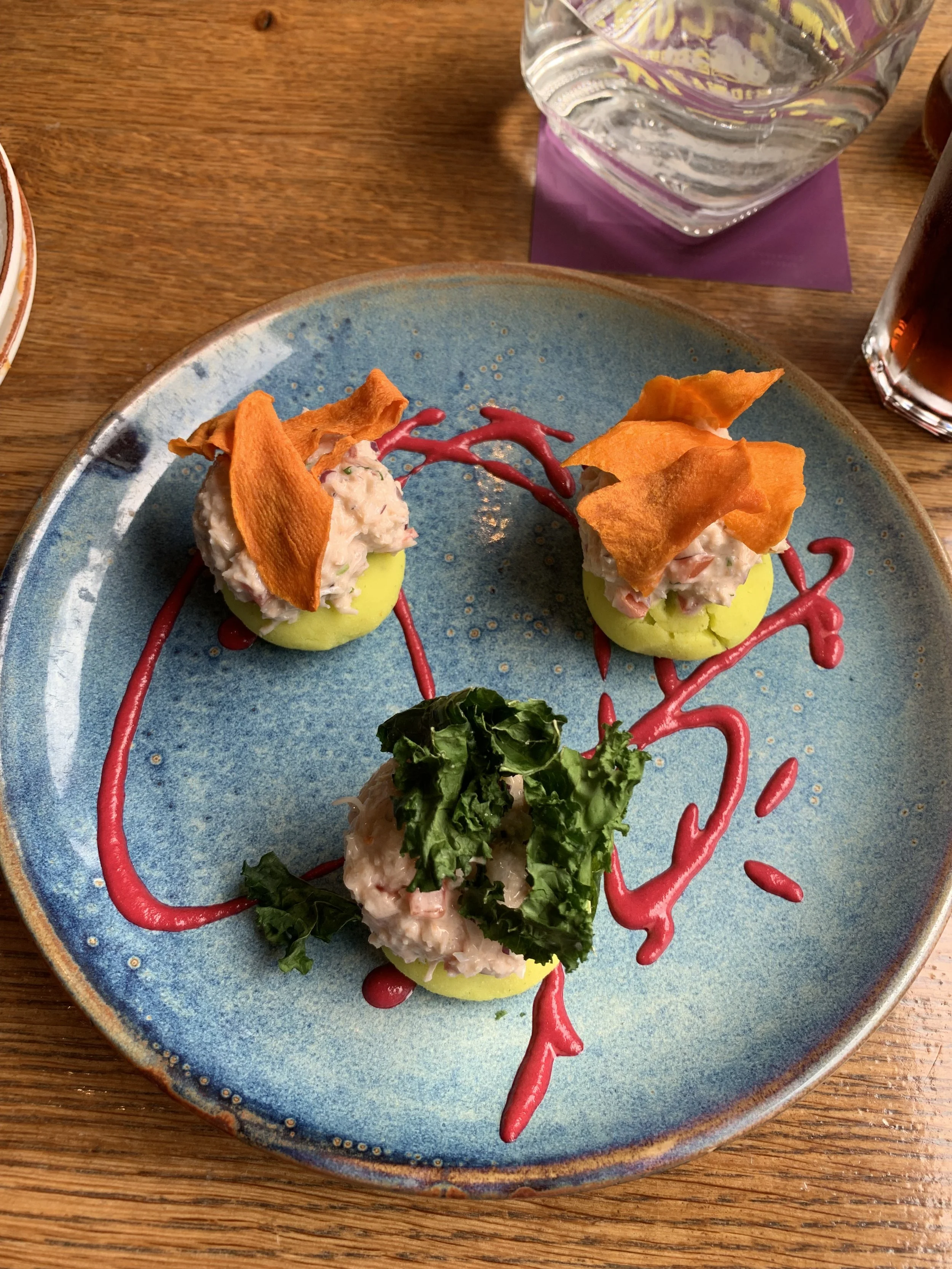 Three appetizers on a blue ceramic plate, each on a round yellow base, garnished with crispy orange chips and leafy greens, with red sauce artfully drizzled on the plate.