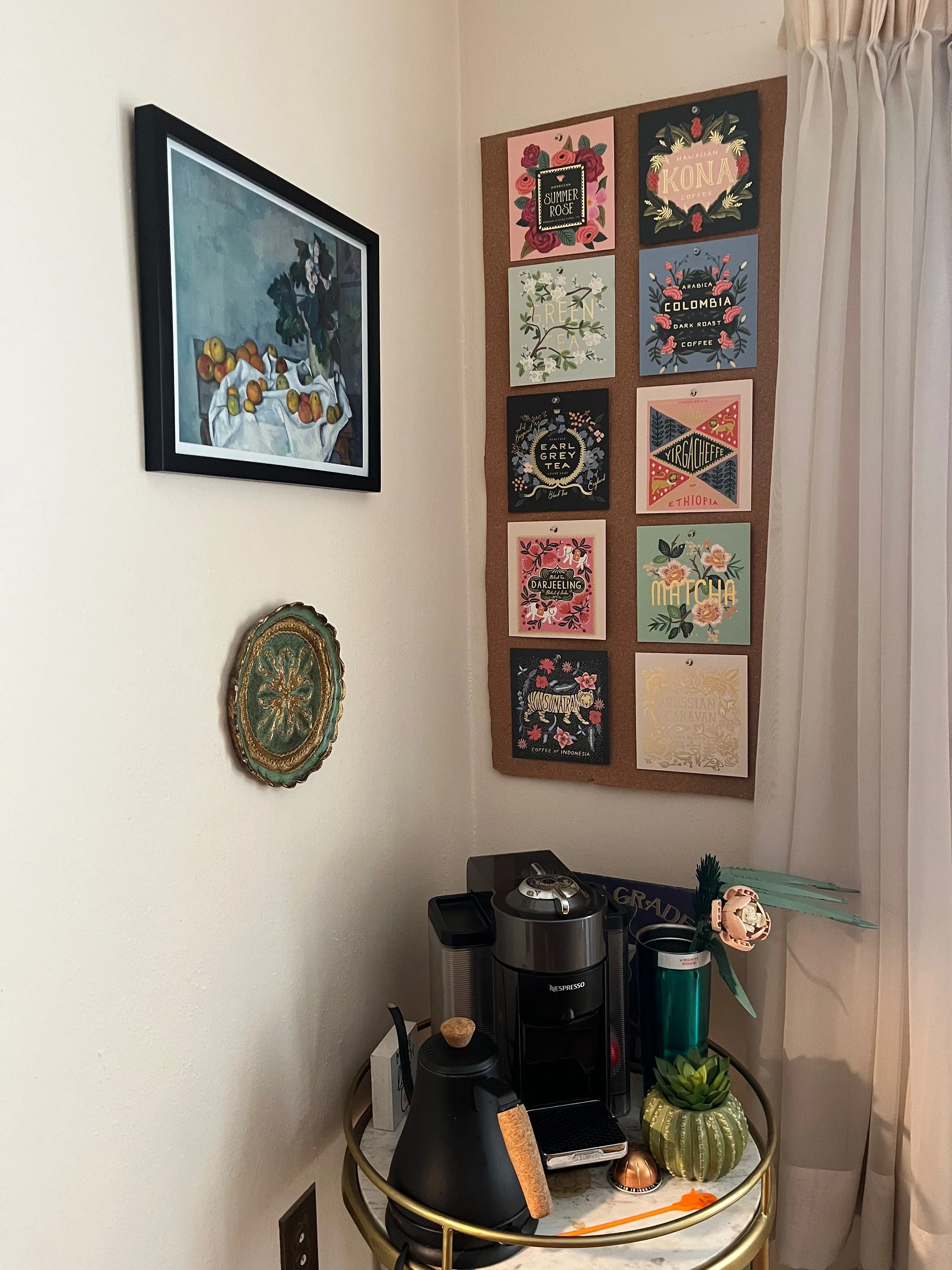 Corner of a room with a small round table holding a coffee machine, a lamp, a potted plant, and a remote control. Behind the table, a corkboard on the wall displays colorful art prints about coffee brands or types. To the left, a wall-mounted paintin