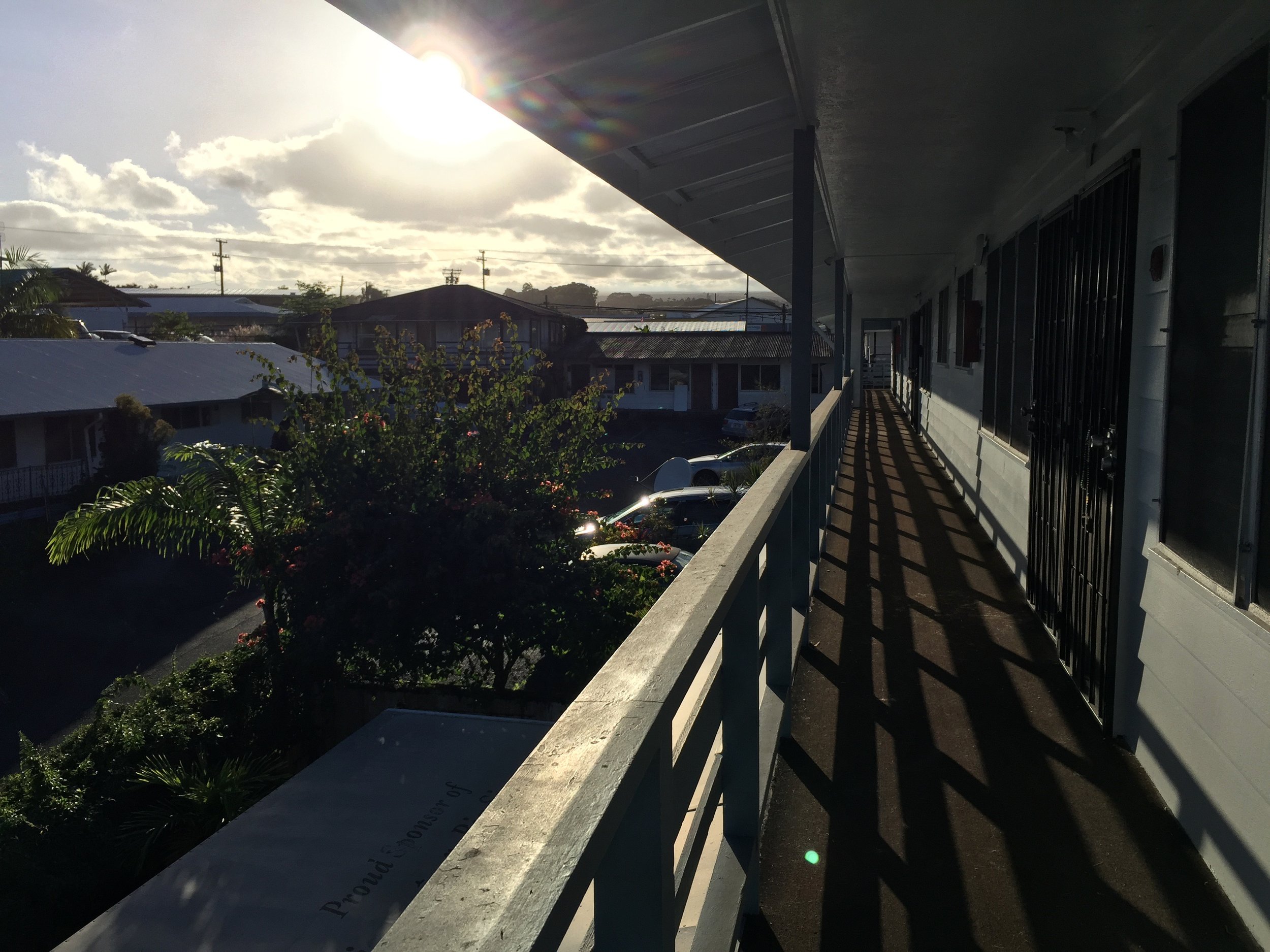 View of an outdoor balcony with houses, trees, and parked cars in the background, illuminated by sunlight with long shadows.