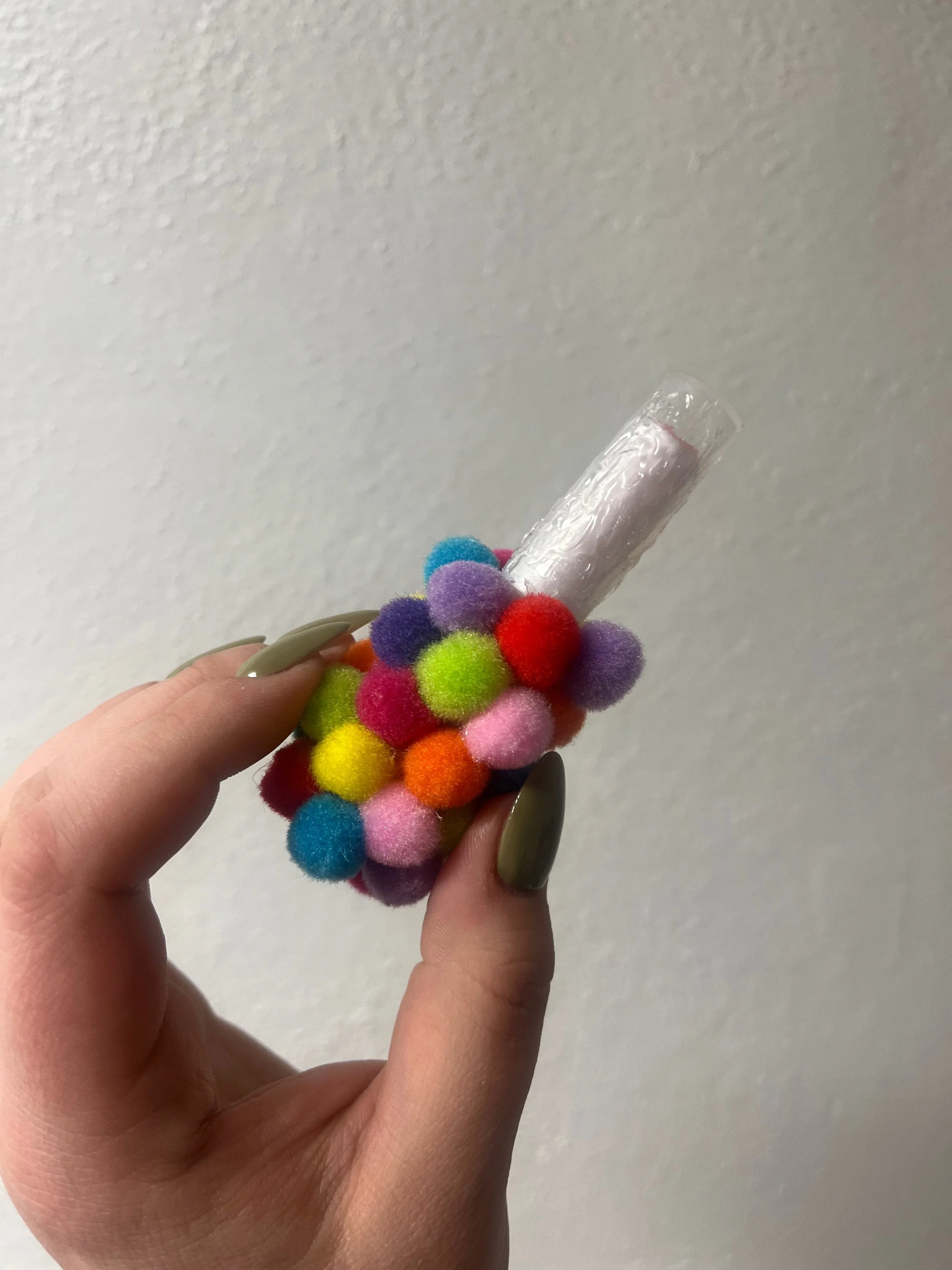 Hand holding a colorful pom-pom decorated in a way resembling a bunch of grapes with a plastic label on top, against a plain wall background.