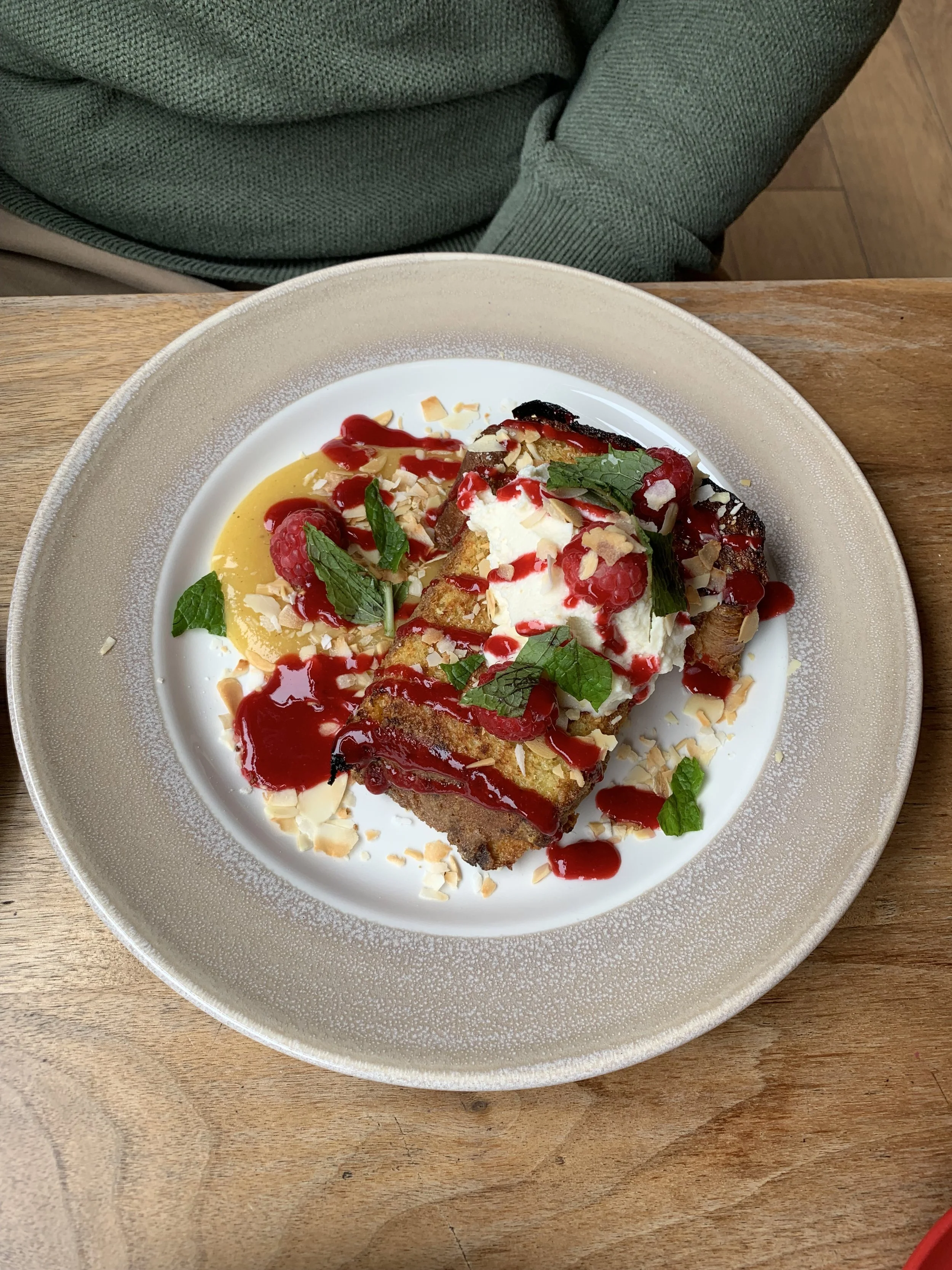 A dessert plate with a slice of French toast topped with whipped cream, raspberries, mint leaves, sliced almonds, and dollops of raspberry sauce on a white and beige plate set on a wooden table.