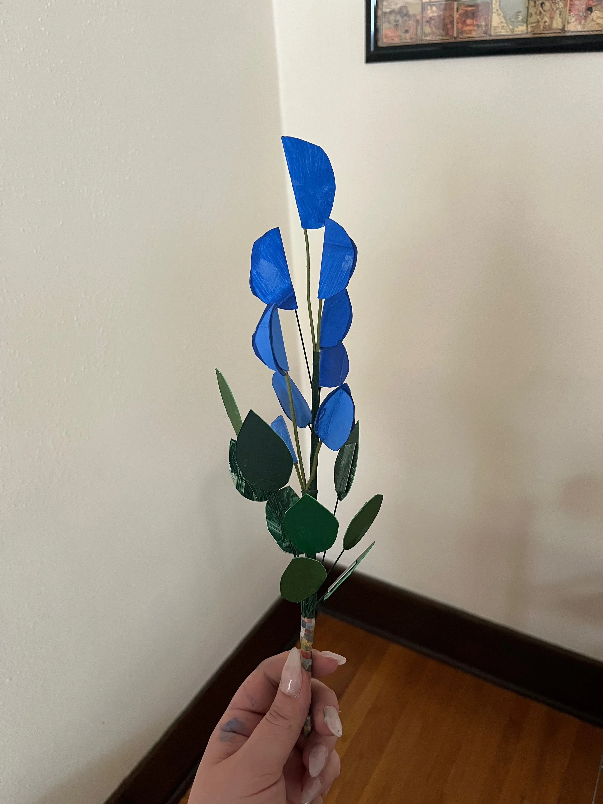 A person holding an artificial flower with blue and green paper petals and leaves, on a paper-wrapped stem, indoors with a wooden floor and a framed picture on the wall.