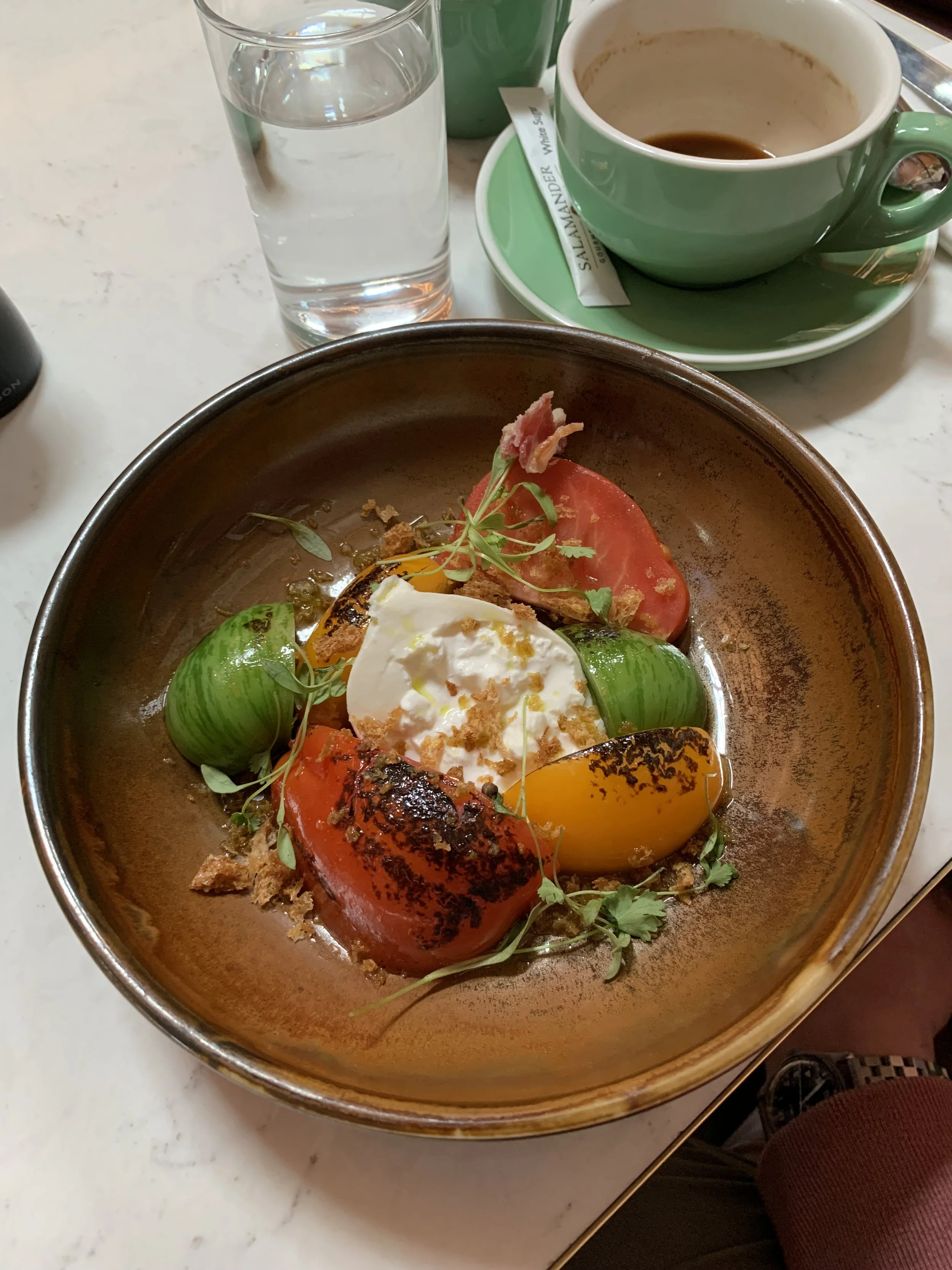 A bowl of roasted vegetables including tomato, zucchini, and bell peppers garnished with microgreens and a poached egg on top, with a glass of water and a cup of coffee in the background.