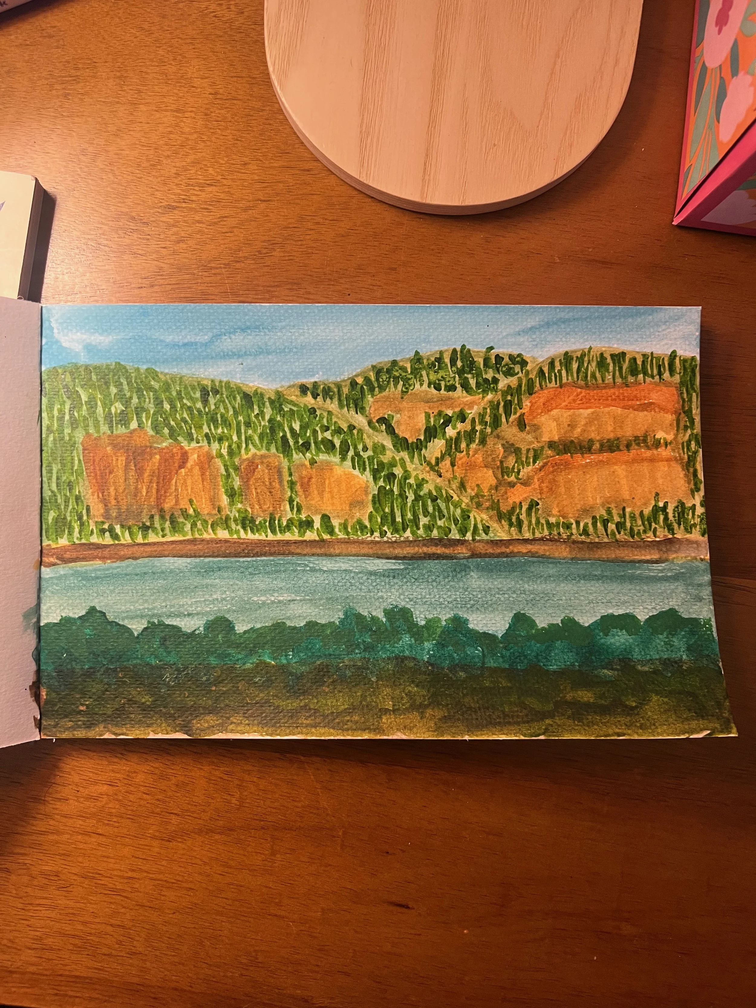 A watercolor painting of a landscape with green hills, orange-brown cliffs, a blue sky, and a body of water in the foreground.