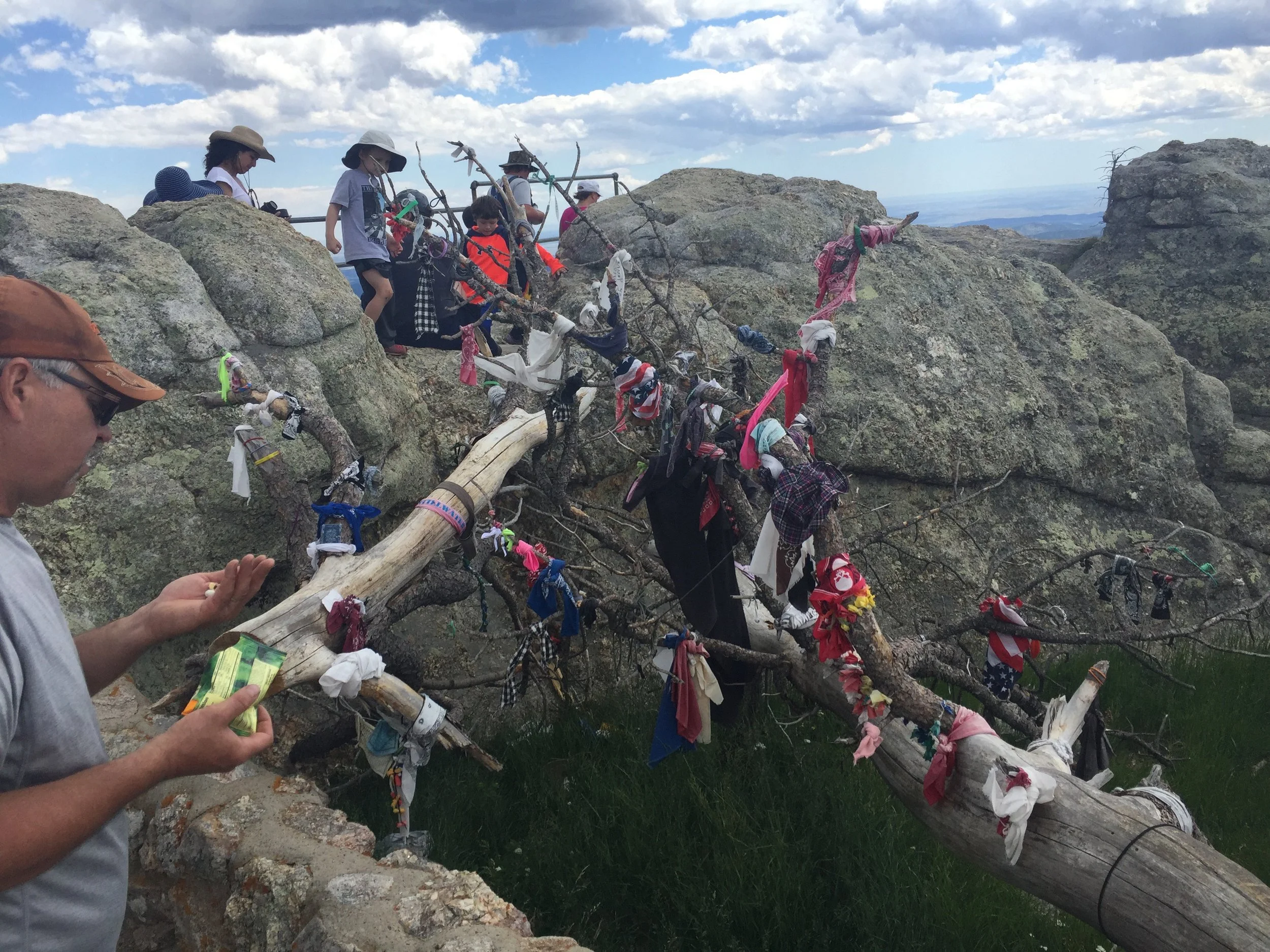 A group of people, including children, sitting and standing on rocks and a fallen tree in a rocky outdoor setting with cloudy sky, with many colorful ribbons tied to the tree branches.