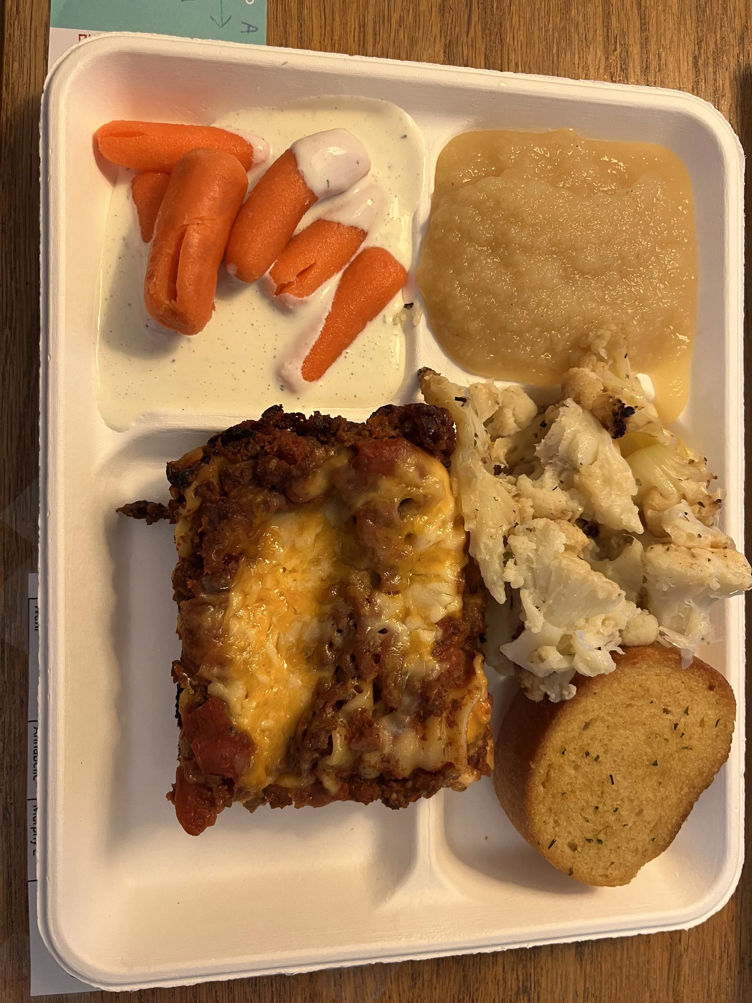 Tray containing carrots with ranch dressing, mashed potatoes, cauliflower with some seasoning, garlic bread, and a serving of lasagna with cheese and meat sauce.