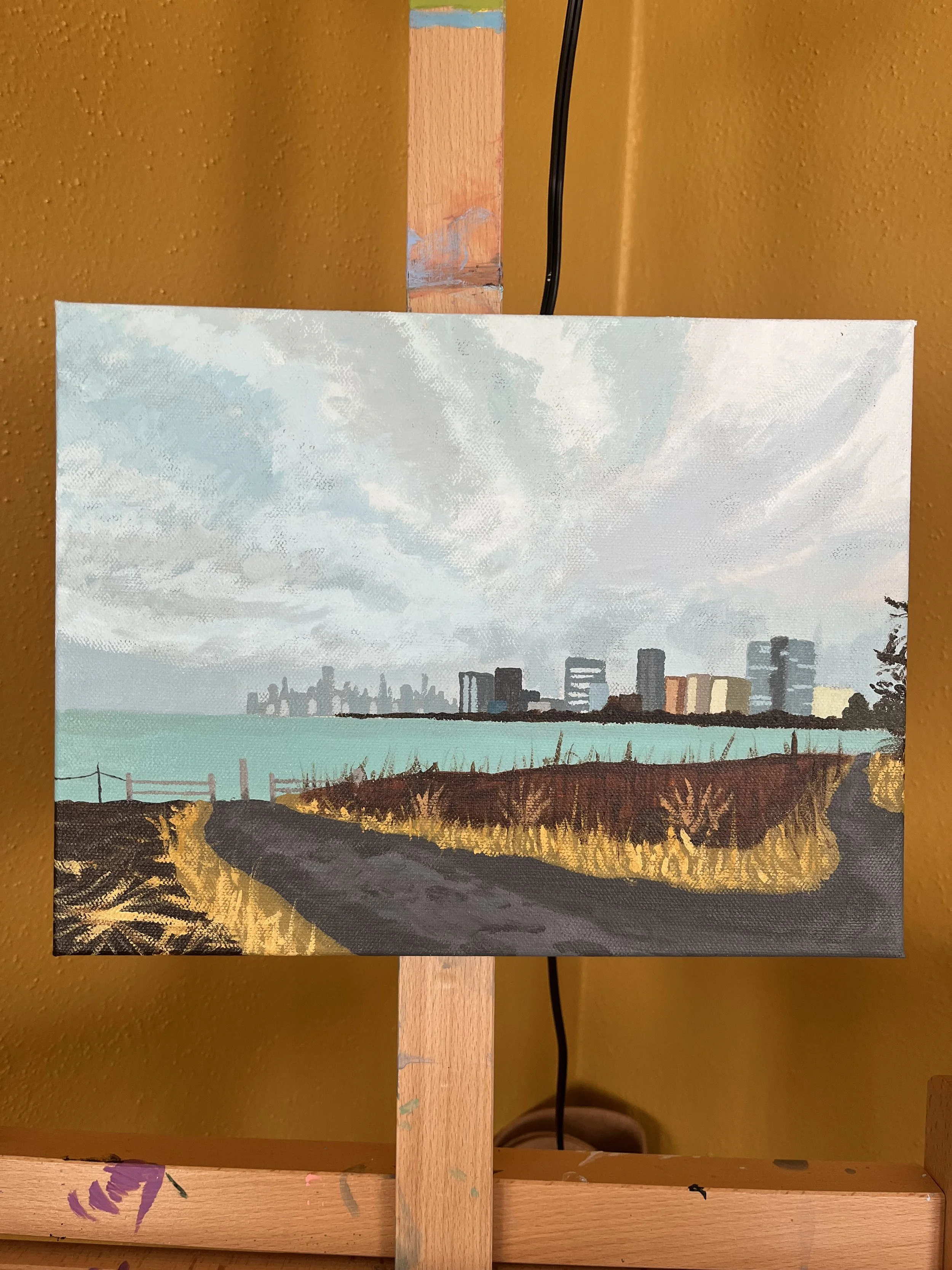 Painting of a city skyline visible from a grassy area with a dirt path, under a cloudy sky.