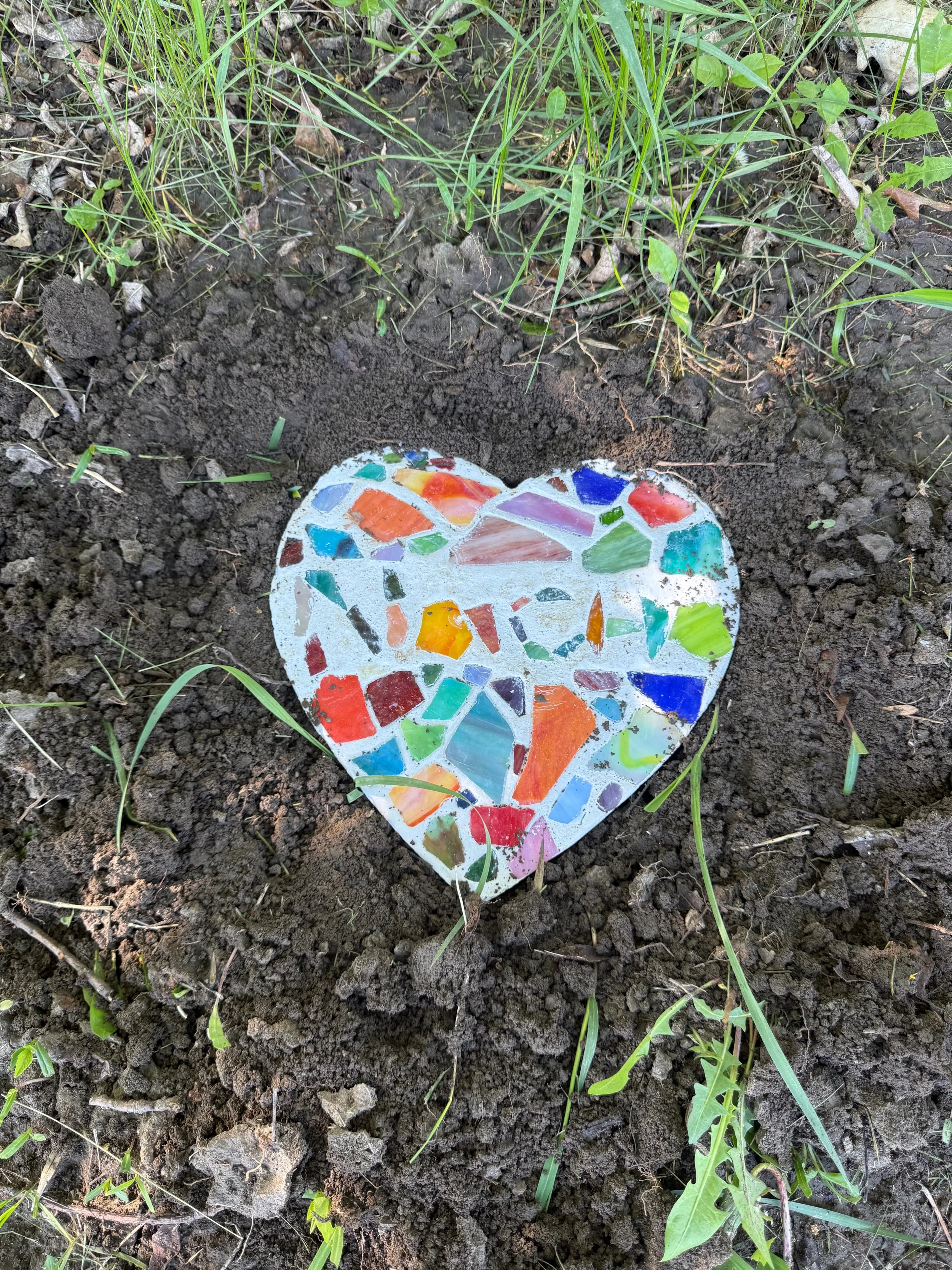 Colorful mosaic heart placed in the dirt surrounded by grass and small plants.