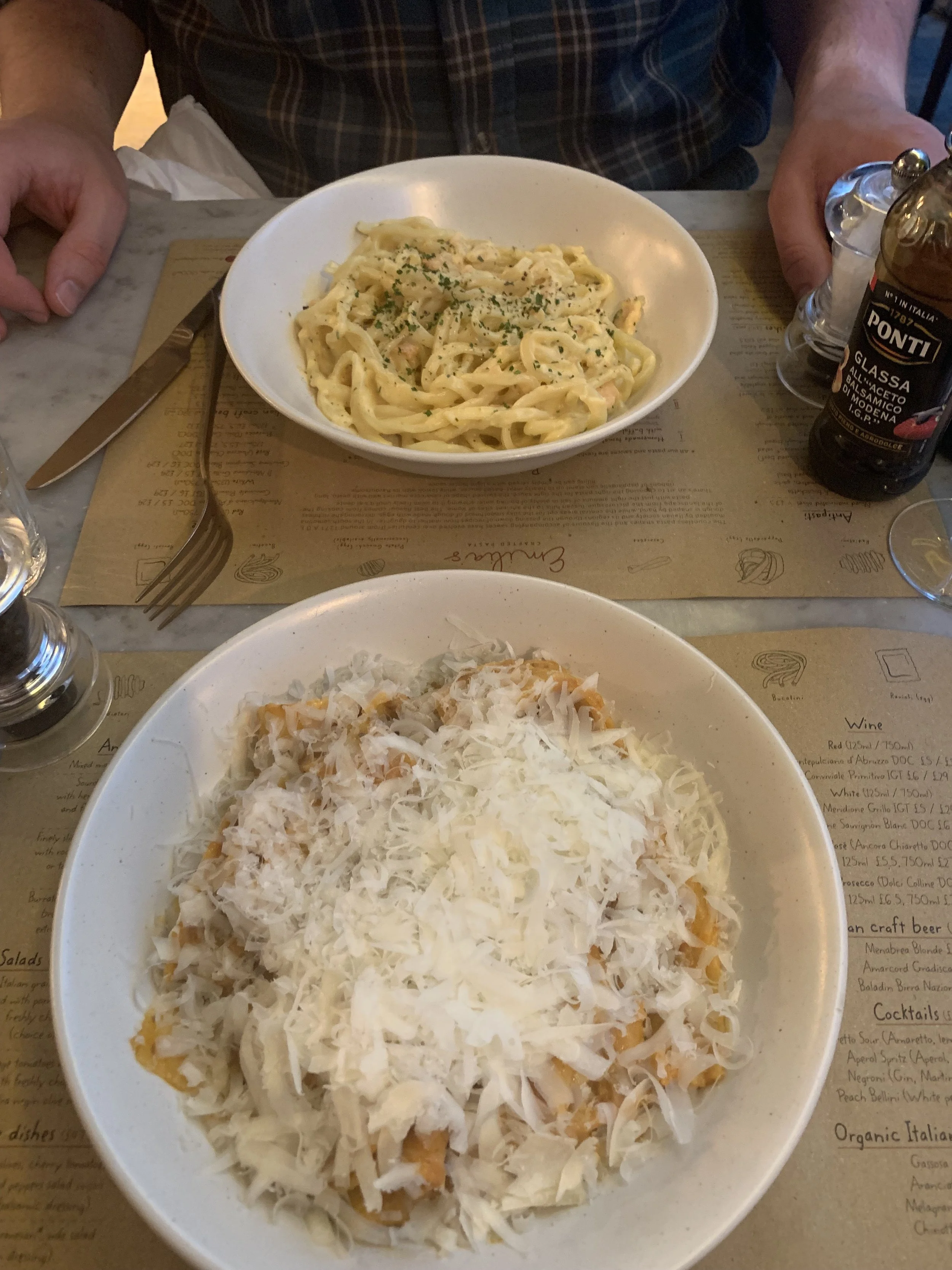 Bowls of creamy pasta and shredded cheese on a restaurant table, with a person's hands nearby and condiment bottles.