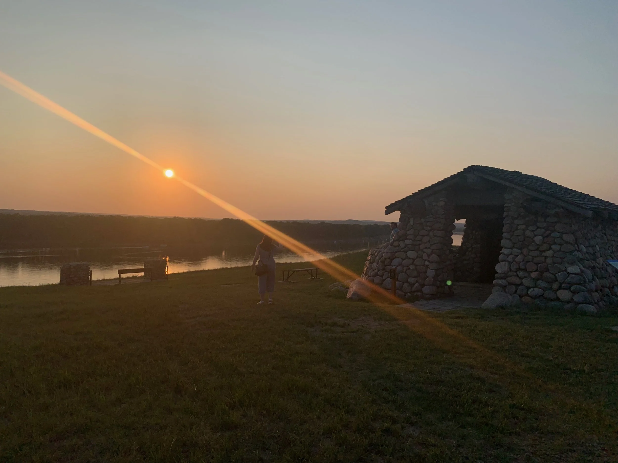 Sunset over a river with a stone cabin in the foreground, two people near the water, and a grassy area.