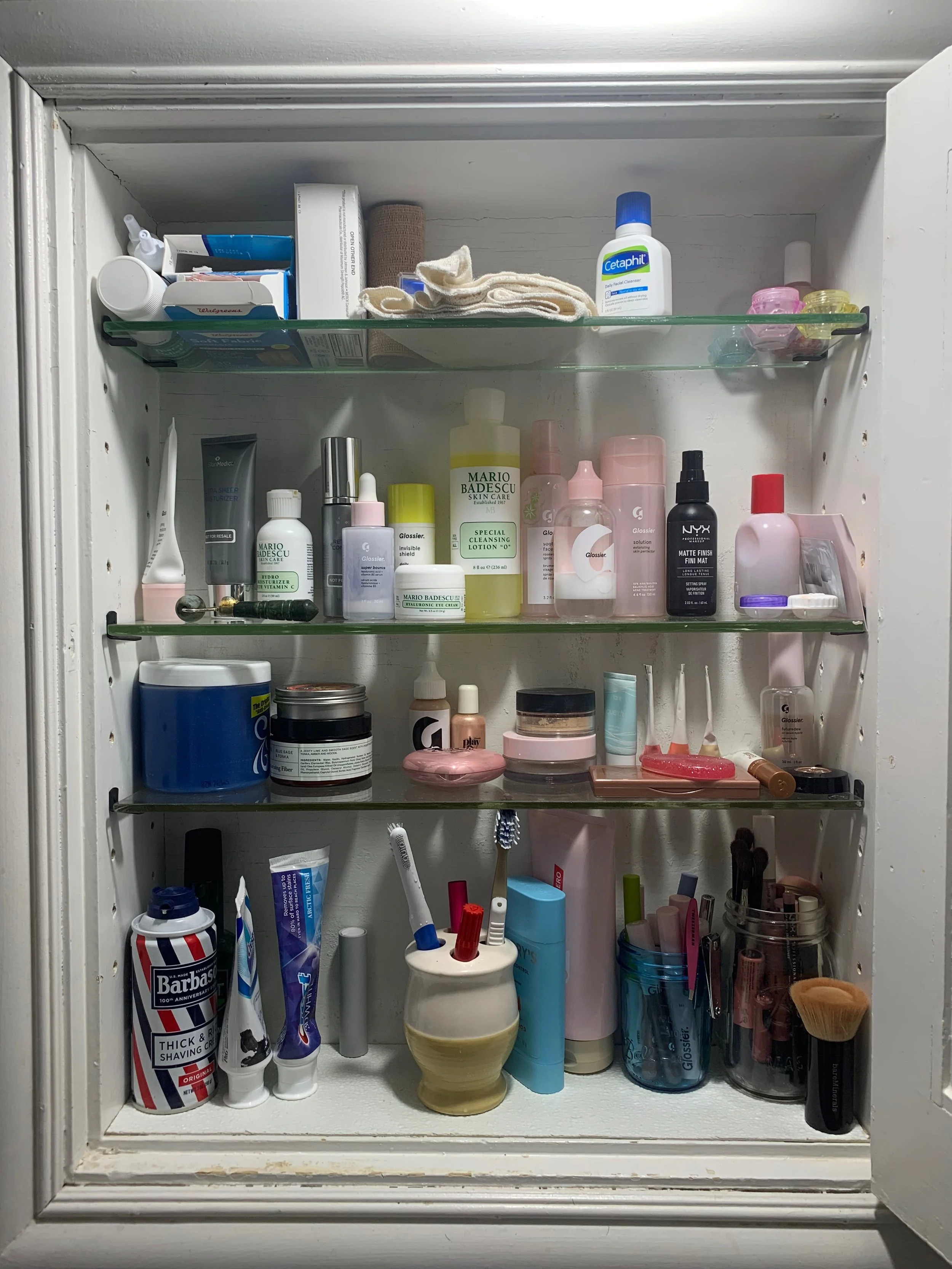 A bathroom cabinet filled with skincare products, toiletries, toothbrushes, and grooming tools organized on four glass shelves.