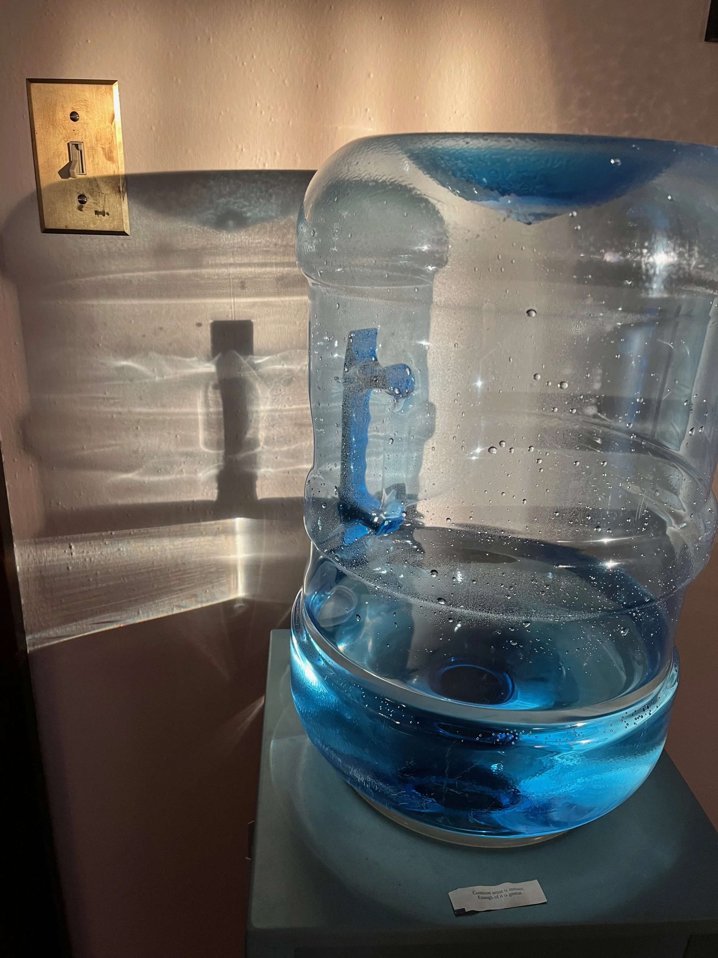 Large transparent water jug filled with water and ice, with a blue plastic spout attached, placed on a pedestal. The background features a beige wall with an electrical outlet and a light switch, casting shadows on the wall.