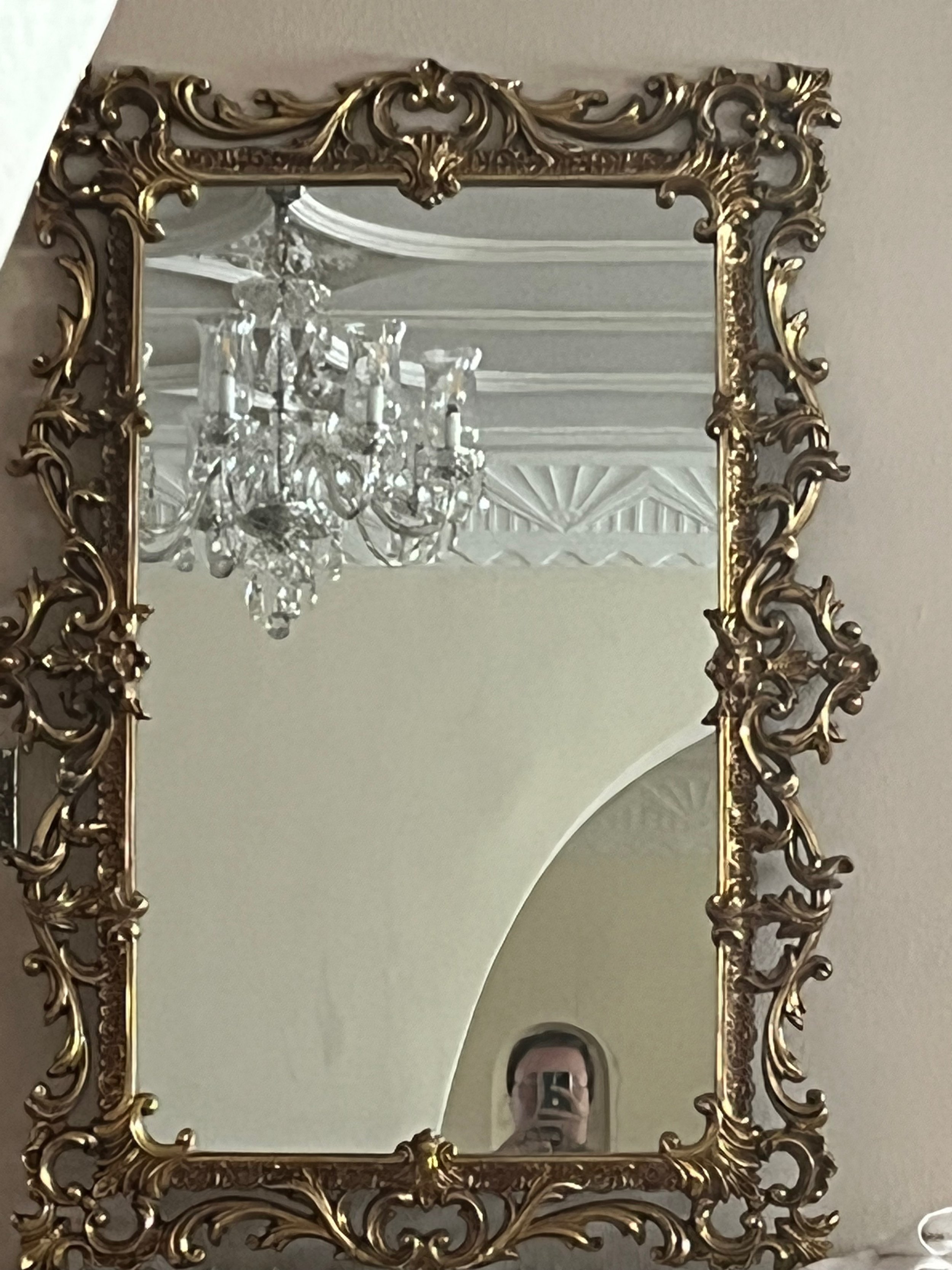 A decorative gold ornate mirror frame reflecting a chandelier and ceiling details, with a person taking a photo visible at the bottom.