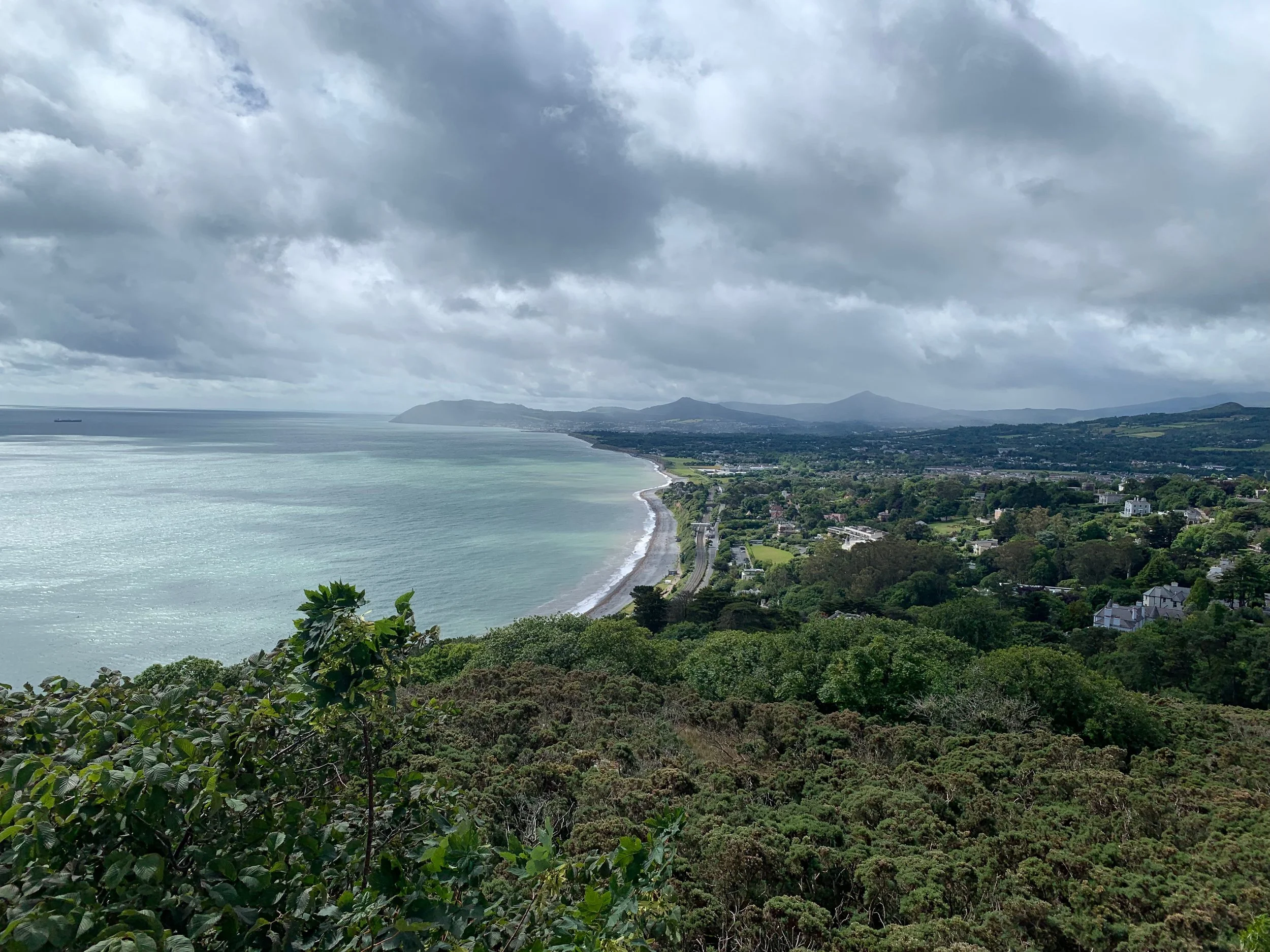 A scenic view of the coastline with sandy beaches, lush green vegetation, and rolling hills under cloudy skies.