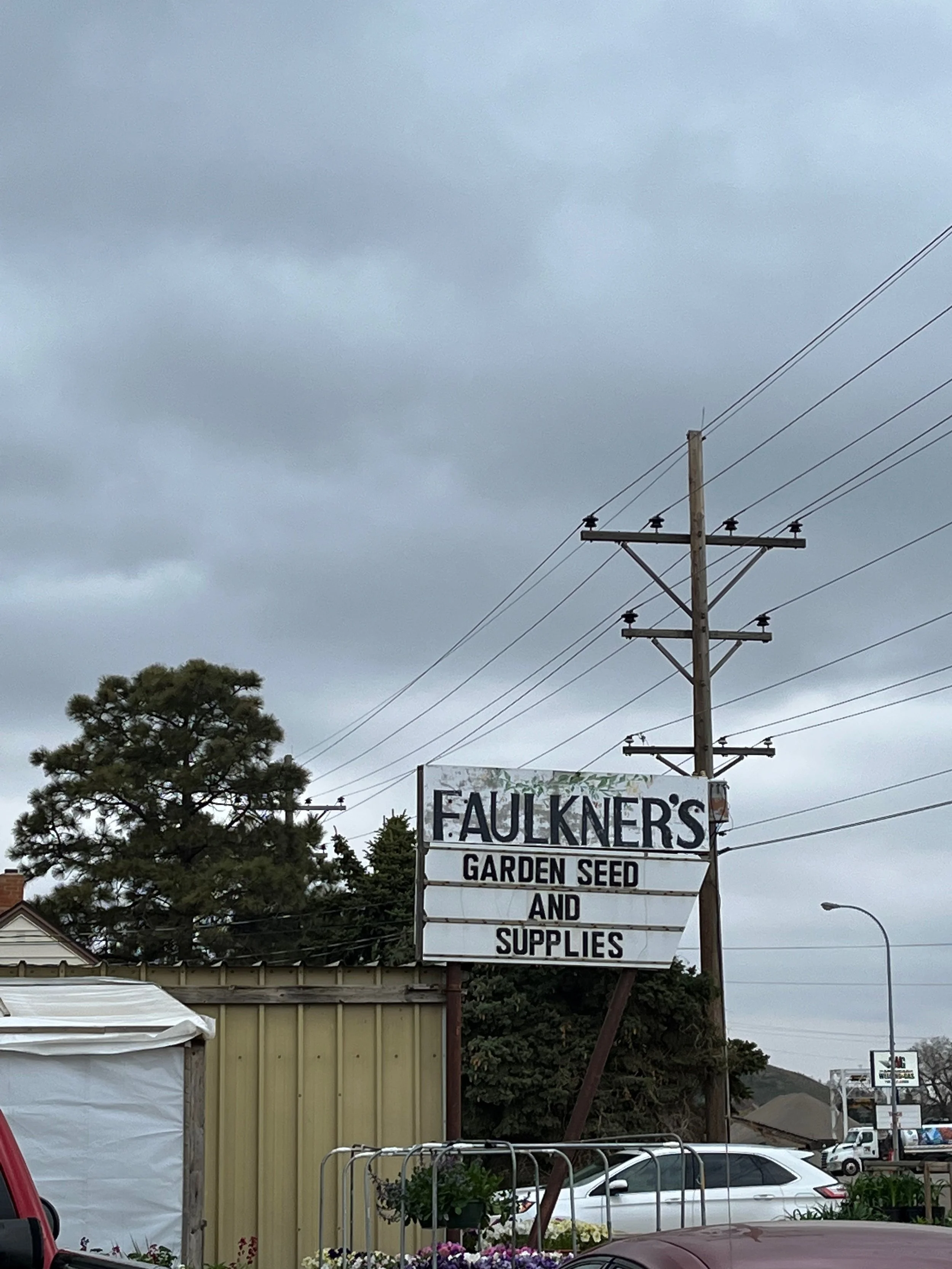 Sign for Faulkner's Garden Seed and Supplies shop, with cloudy sky and cars parked nearby.