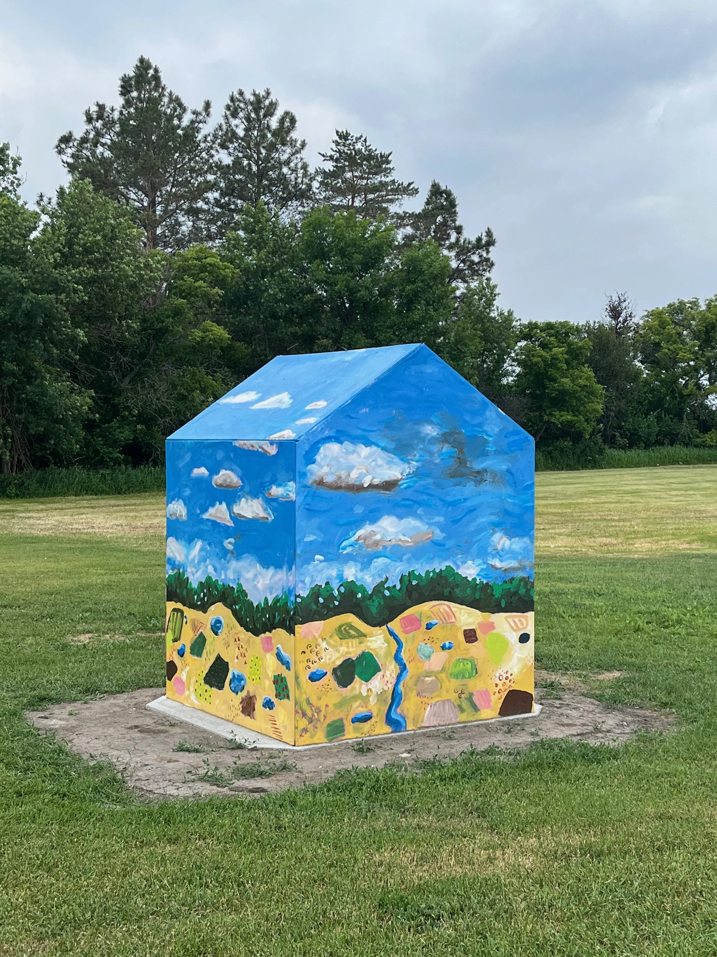 A small rectangular building or box painted with a landscape scene, including a bright blue sky with clouds, a line of trees, a yellow field with various abstract shapes, and a blue stream running through the field, located in an open grassy area wit
