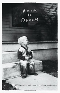 Black and white photo of a young boy sitting on a step outside a house, looking up at a window with the words 'Room to Dream' written on it, with trees and a house in the background.