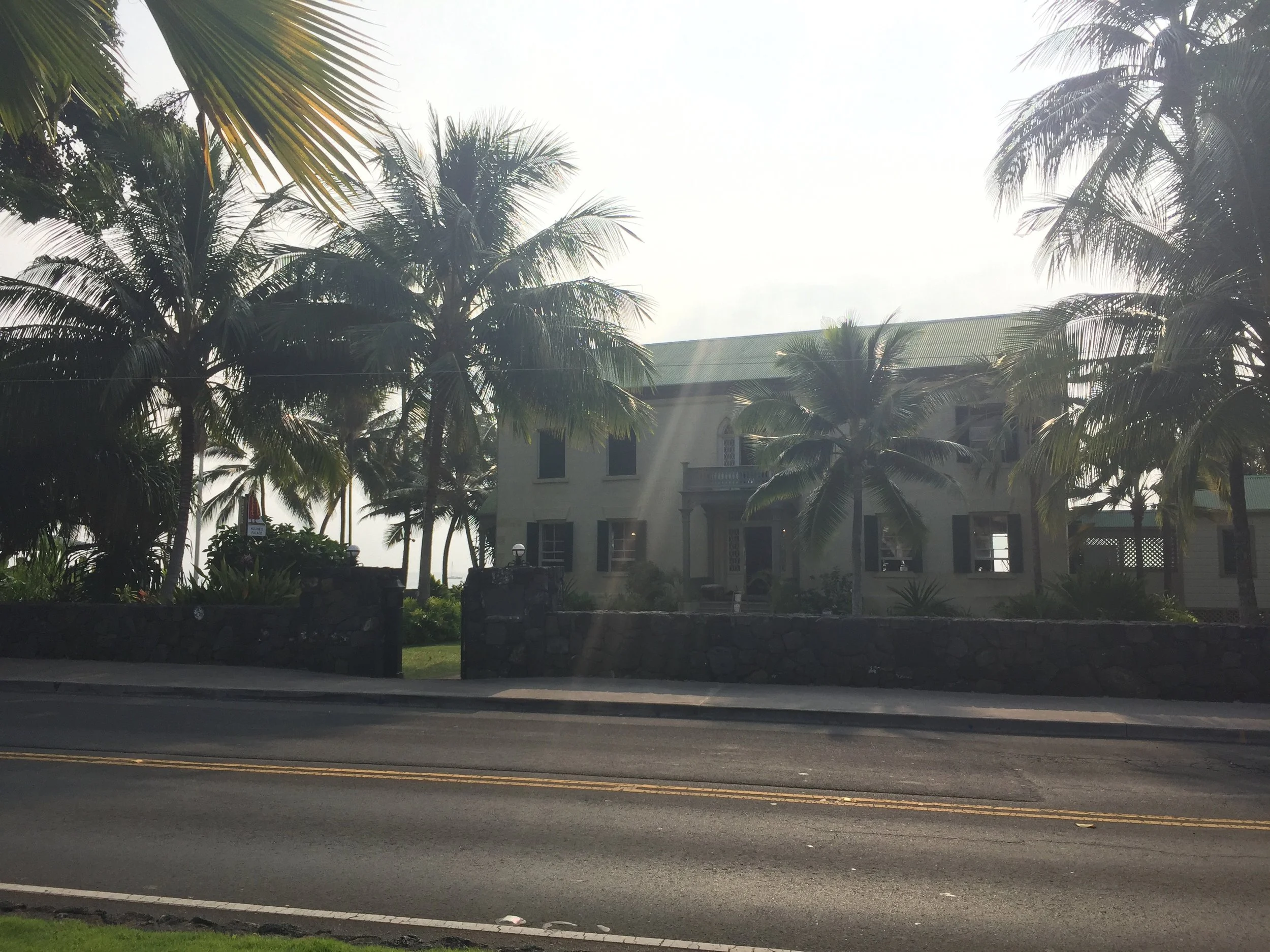 A large house behind a stone wall with a driveway, surrounded by tall palm trees in a sunny setting.