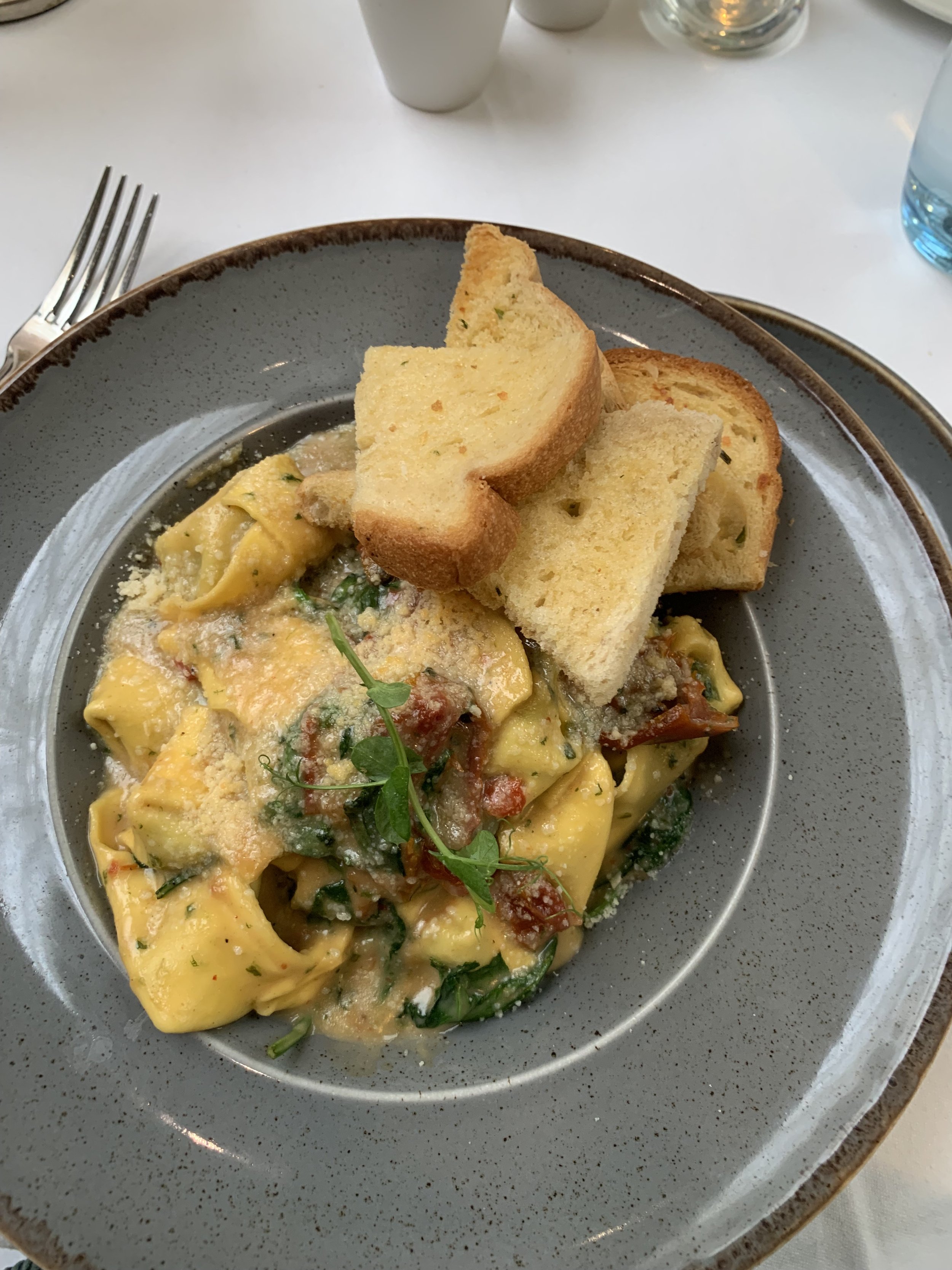 Plate of egg pasta with vegetables and cheese, garnished with microgreens, served with toasted bread slices on a gray plate.