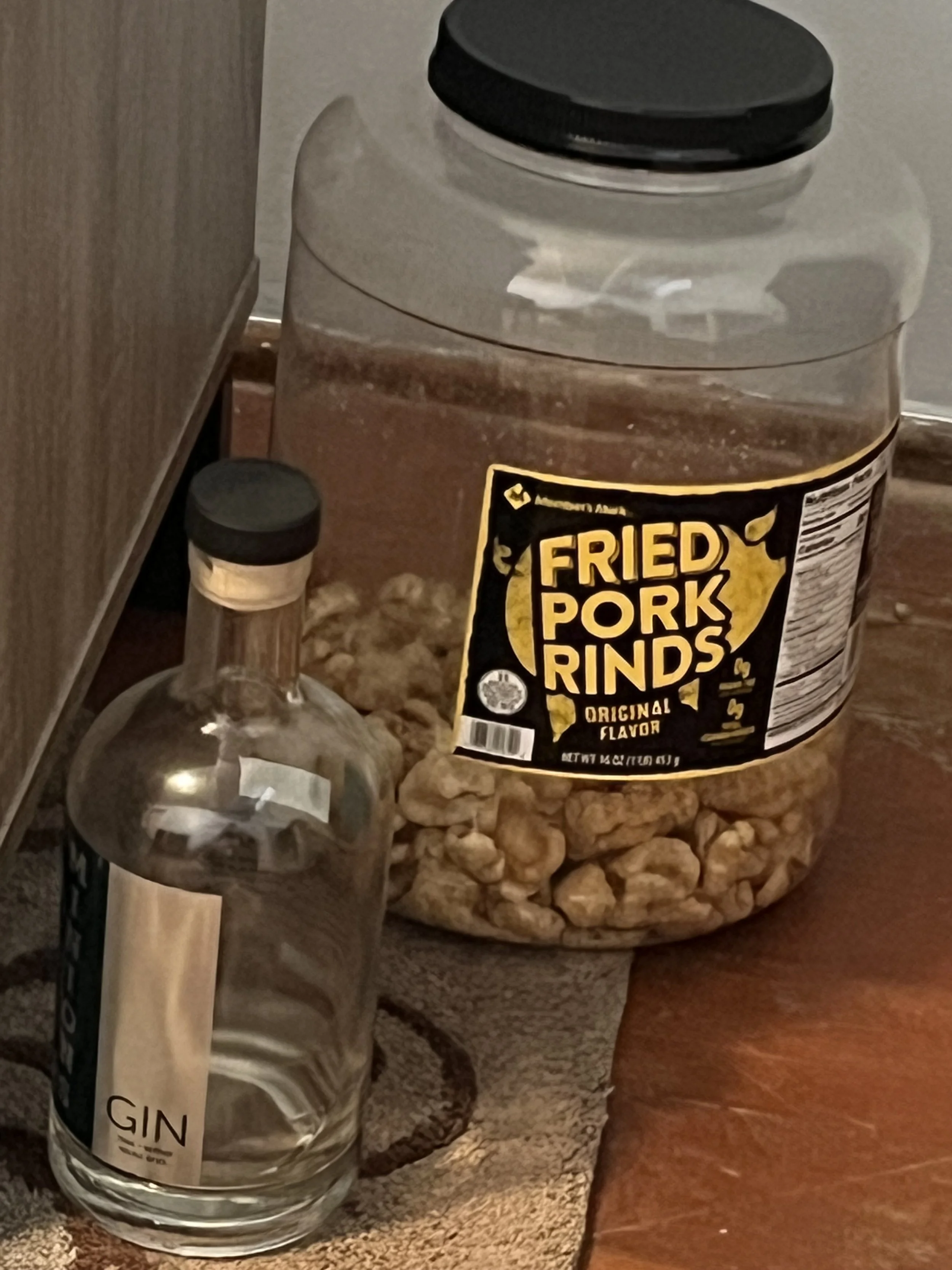 A jar of fried pork rinds and a small clear bottle of gin on a wooden surface.
