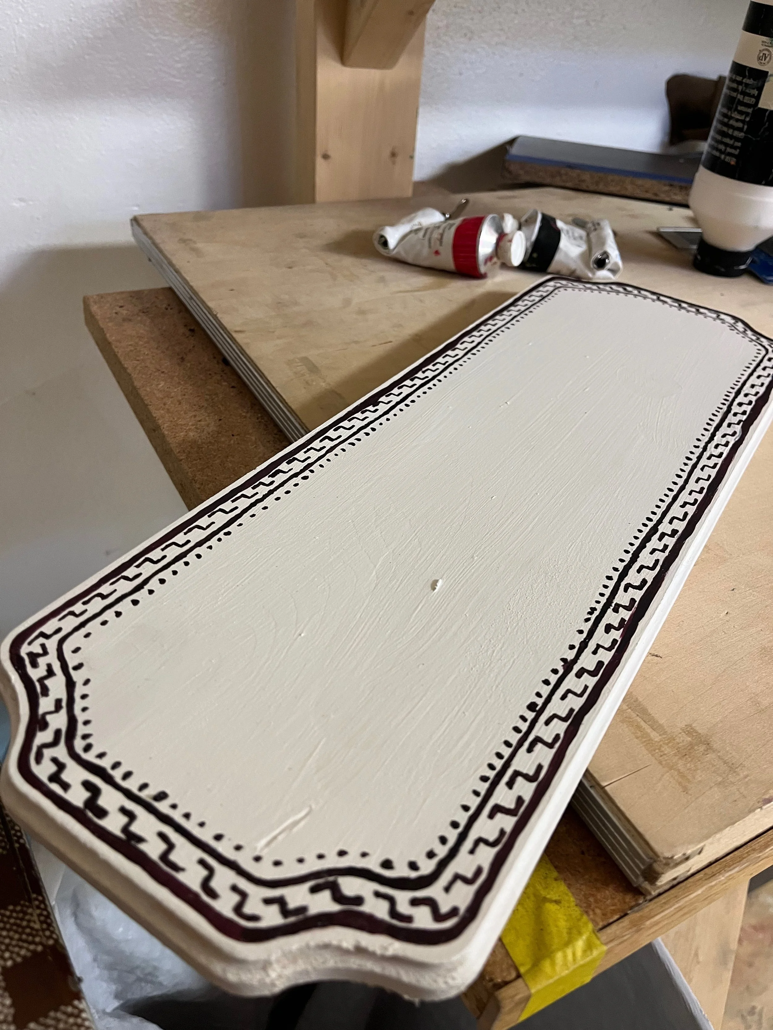 A painted decorative wooden sign with a white background and black border design, resting on a workbench in a workshop.