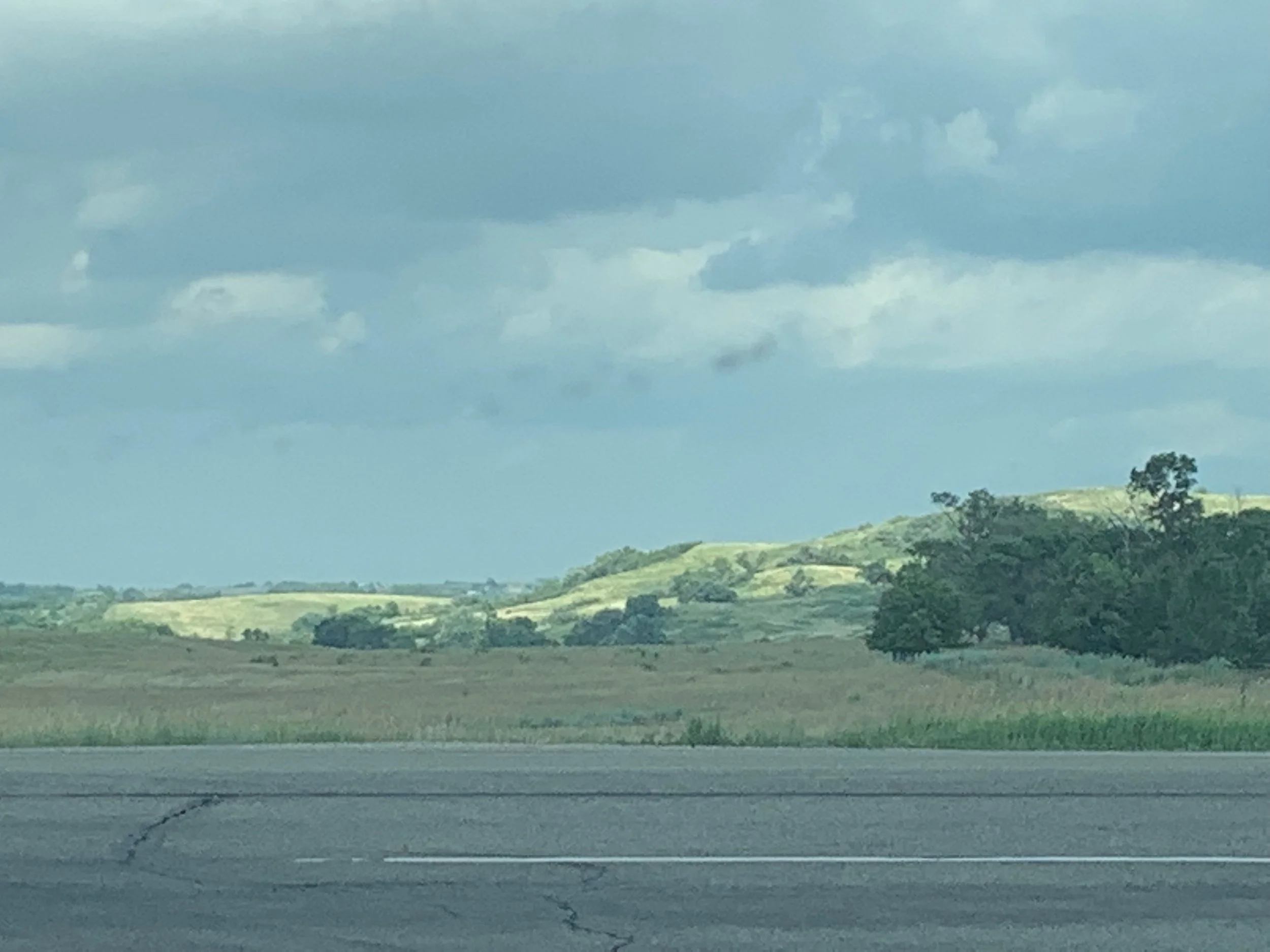 Scenic view of rolling green hills with a cloudy sky, seen from a road in the foreground.