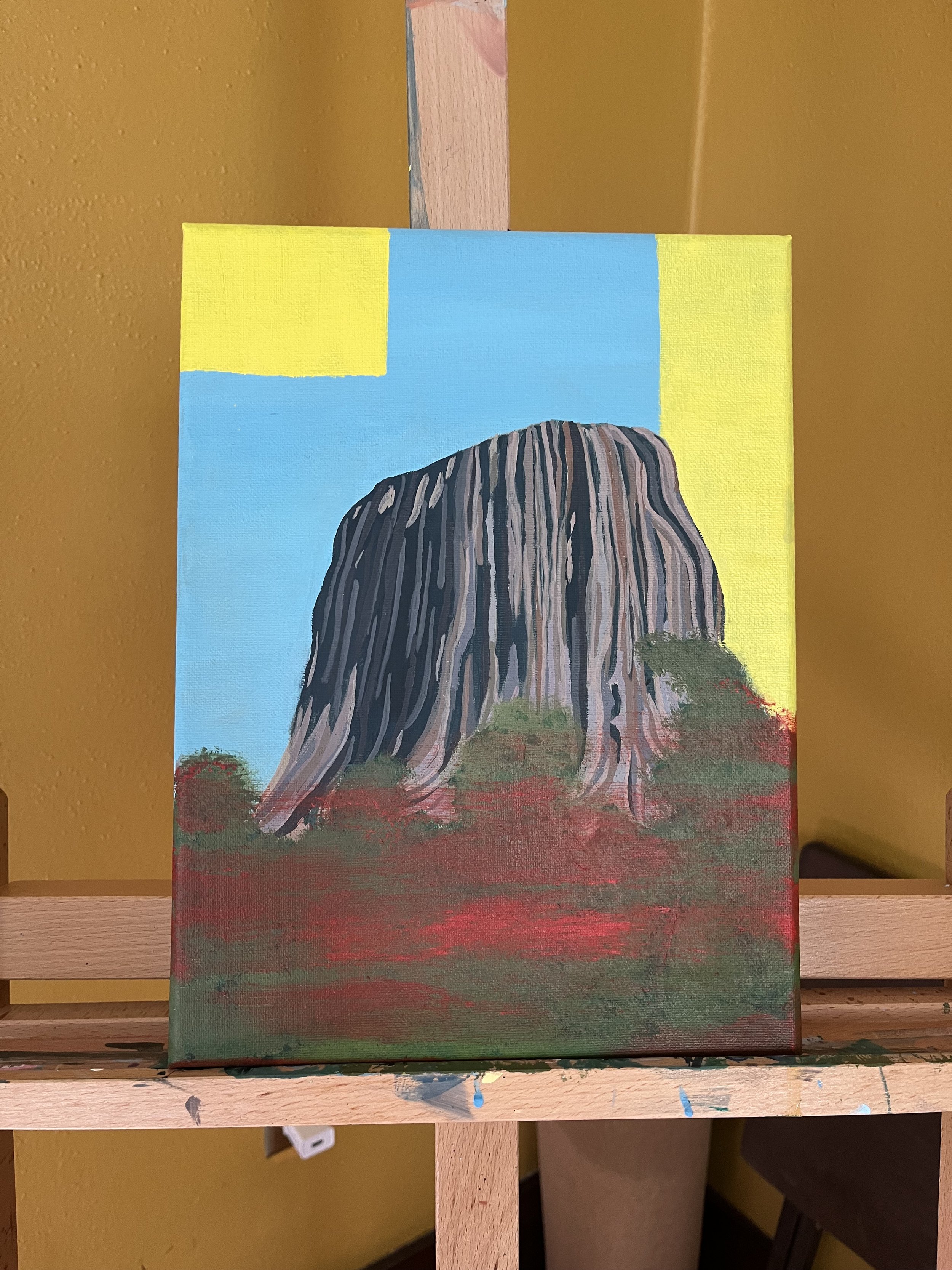 Painting of a large mountain with vertical striped patterns in grayscale, set against a blue sky with a yellow patch and a green and red landscape at the base.