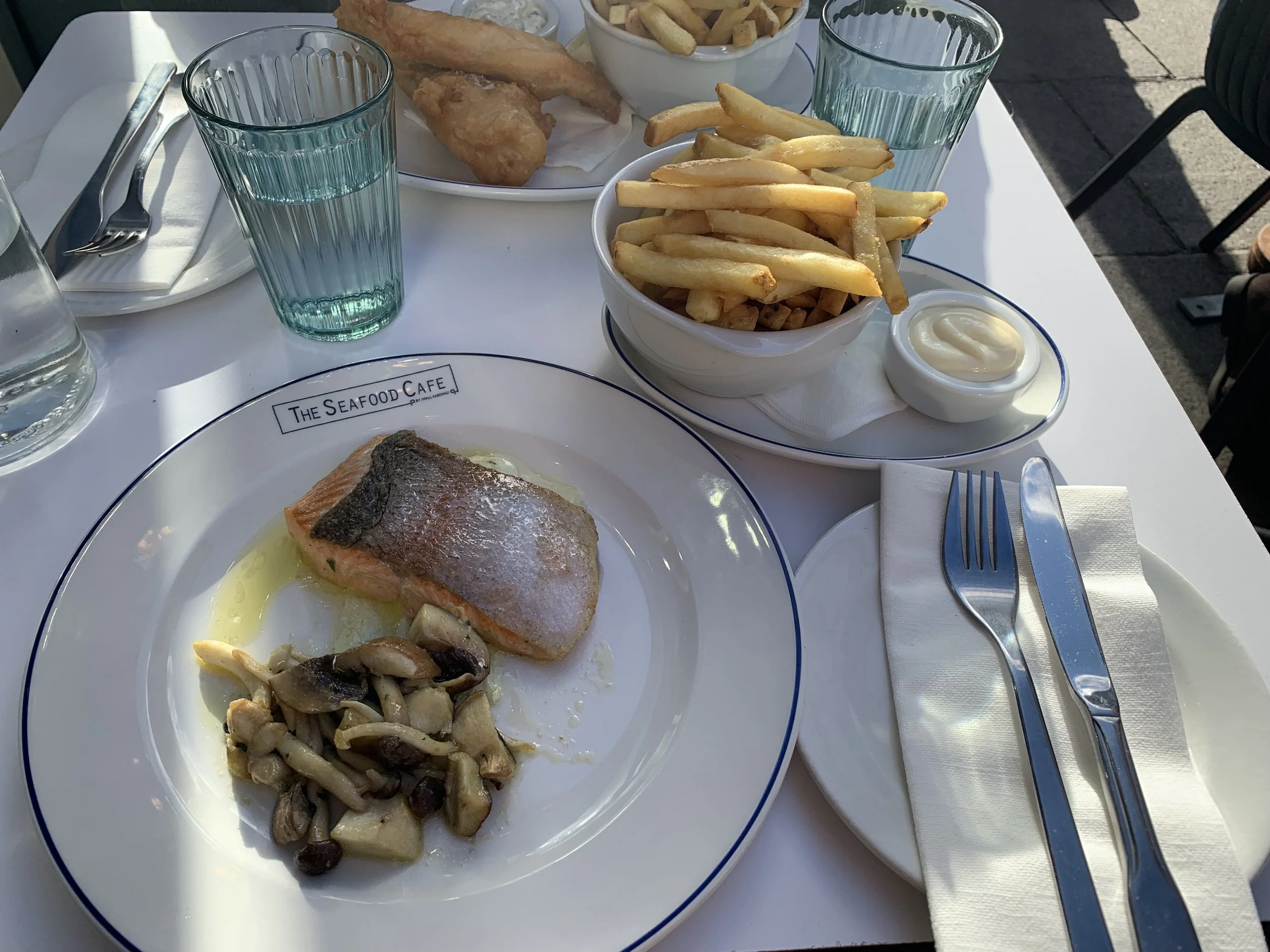 A plate with a cooked salmon fillet topped with a slice of crispy skin, served with sautéed mushrooms. Also visible are bowls of French fries, a small dish of mayonnaise, a glass of water, and cups of water on a white table at The Seafood Cafe.