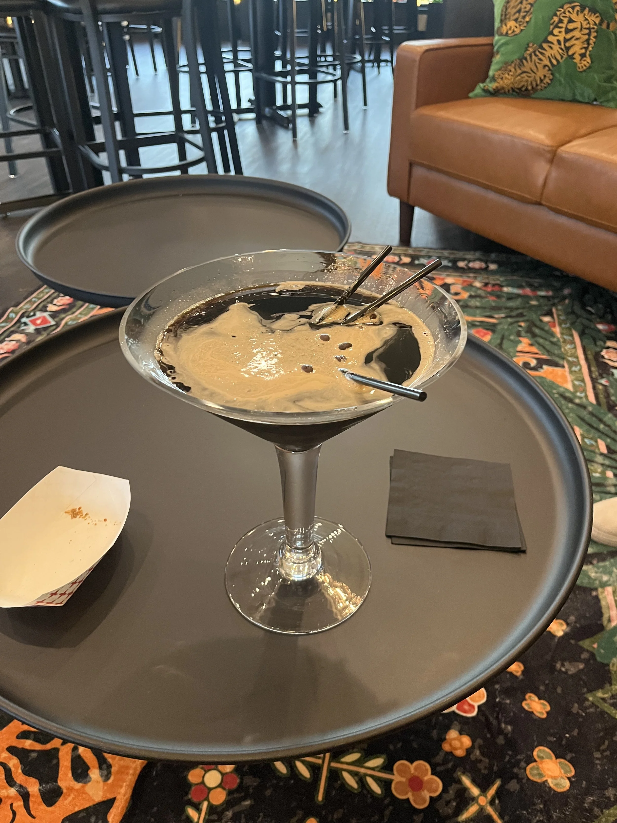 A cocktail glass with dark coffee-colored liquid topped with foam, placed on a round black tray on a table.