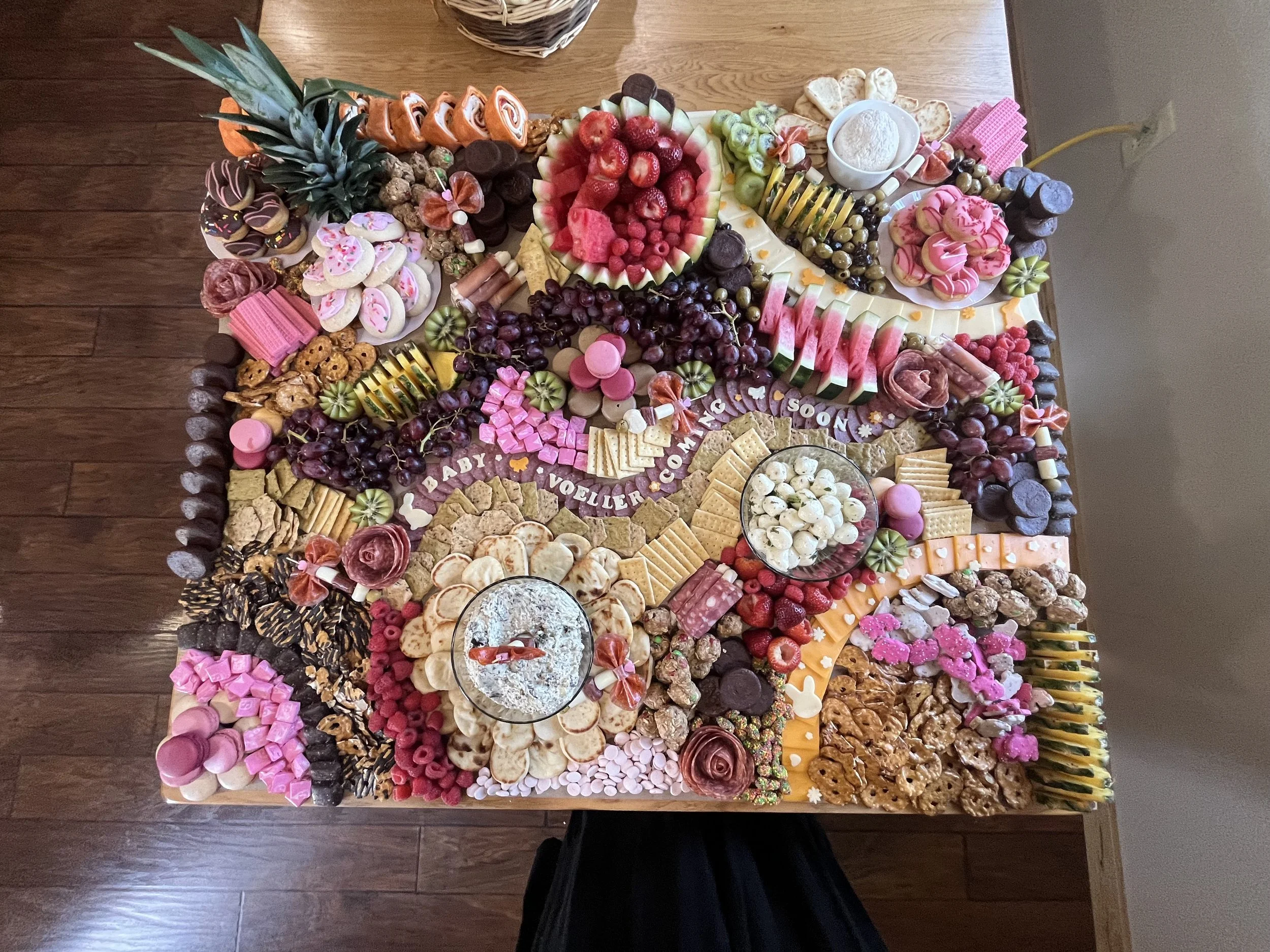 A colorful assortment of various candies, chocolates, fruits, and baked goods arranged on a large table, including watermelon, strawberries, grapes, cookies, marshmallows, and chocolates, with a message that says "Baby Vouiller Coming Soon."