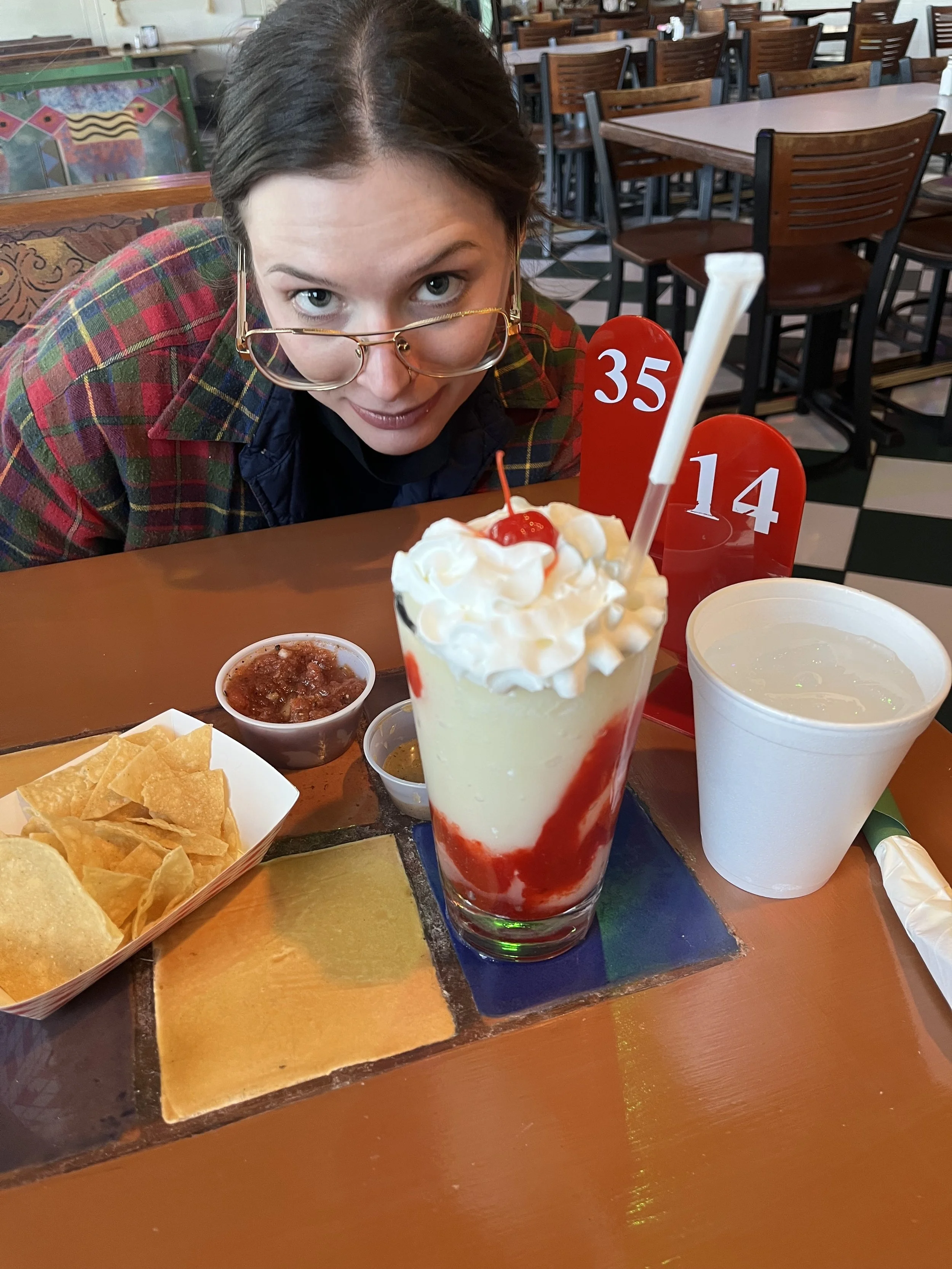 A woman with glasses leaning over a table in a restaurant, looking at a large milkshake topped with whipped cream and a cherry, with tortilla chips, salsa, and a drink on the table.