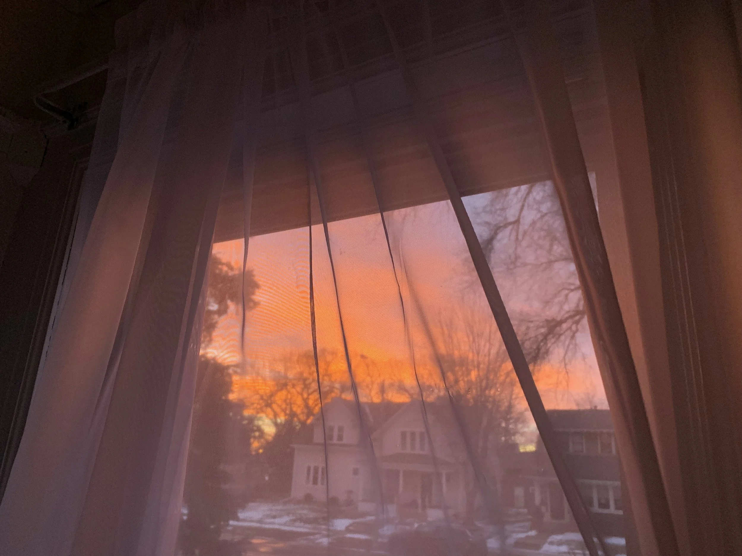 Sunset sky with orange and pink hues viewed through a window with sheer pink curtains and blinds.