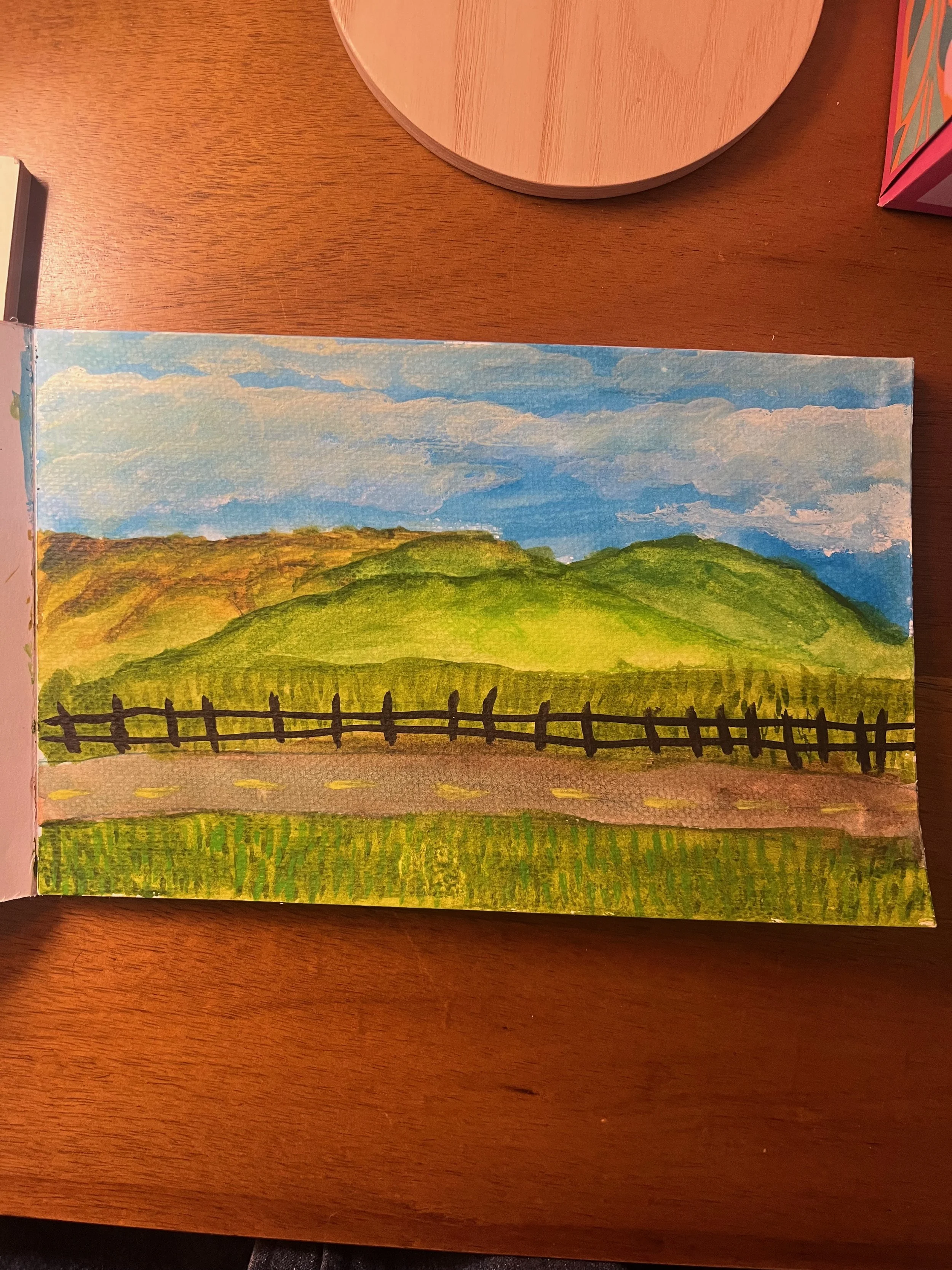 A watercolor landscape painting of green hills under a blue sky with white clouds, featuring a wooden fence in the foreground and a road at the base of the hills.