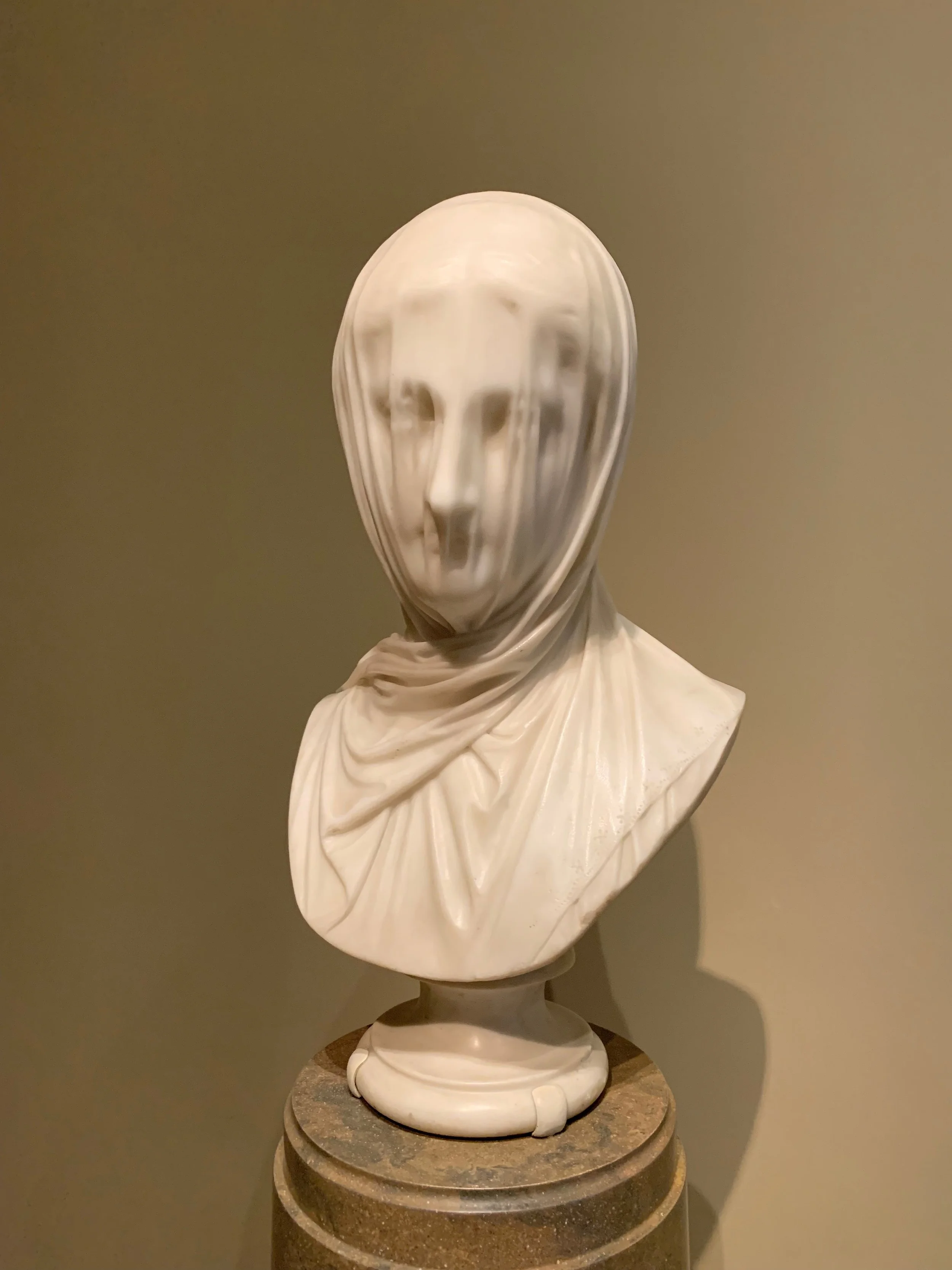 Marble bust sculpture of a woman with a hood, slightly distorted face, placed on a round pedestal.