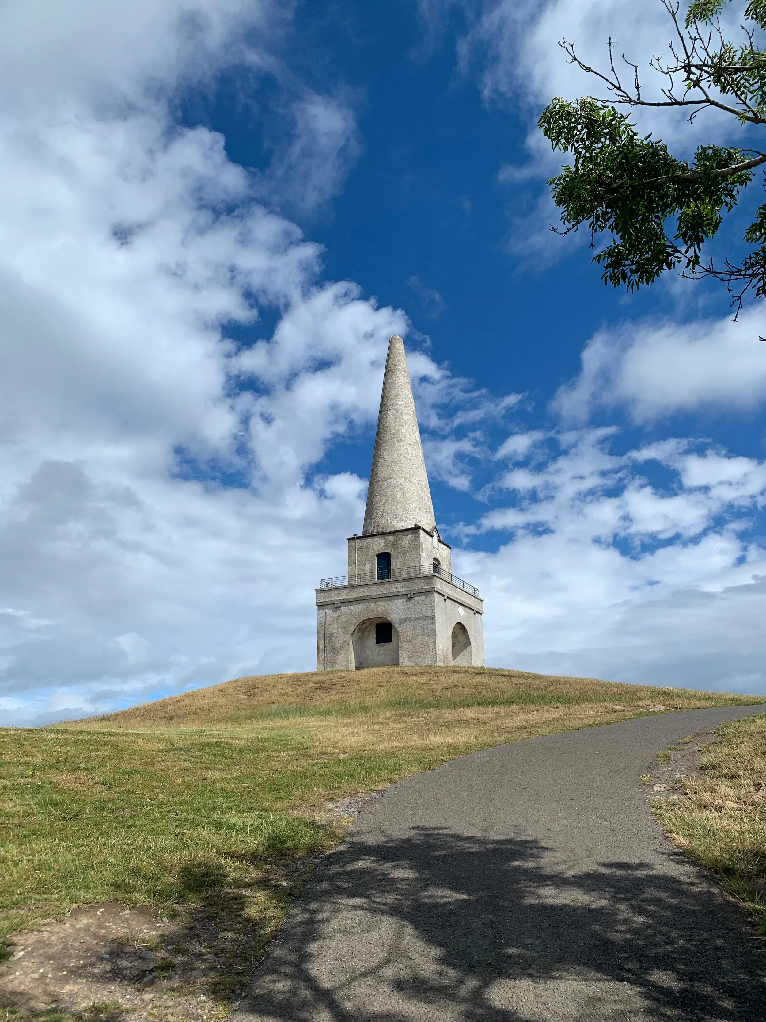 A tall, narrow stone tower with a conical top on a grassy hill, with a pathway leading up to it, under a partly cloudy sky.