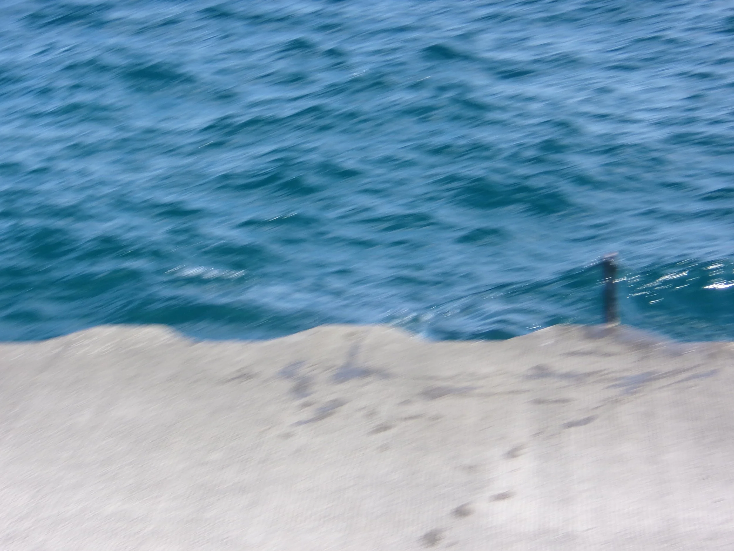 Blurred image of ocean water near a sandy shoreline with a small submerged dark object or marine structure near the edge.