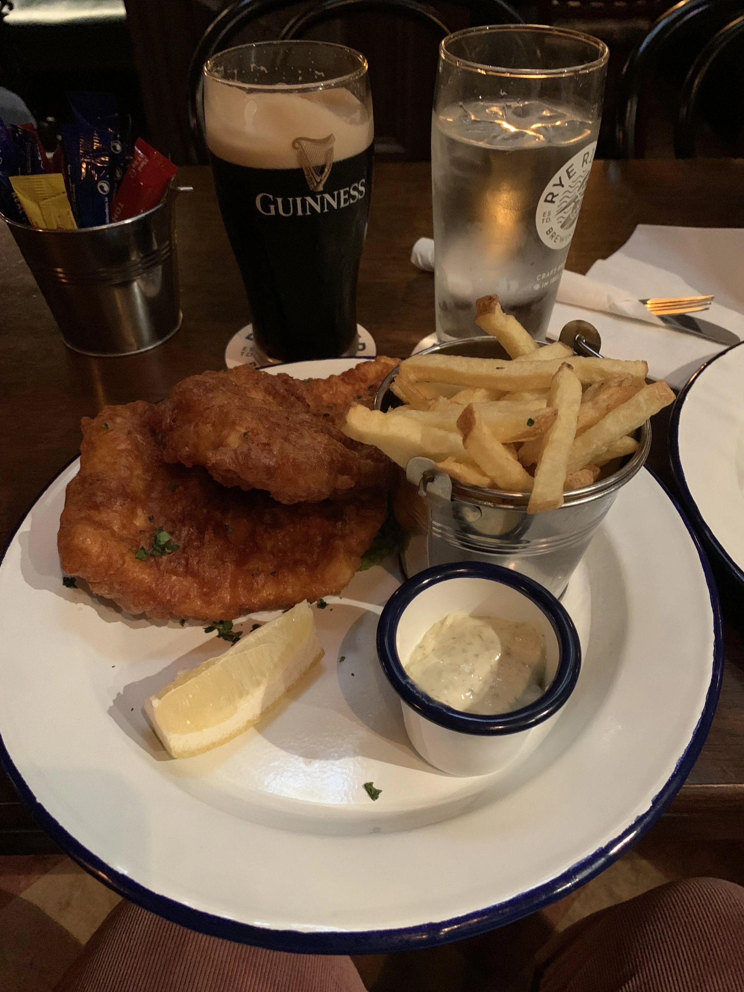 A plate of fried fish and chips with tartar sauce, lemon wedge, and a side of French fries, with a glass of Guinness beer, a glass of water, and condiments on a dark wooden table.