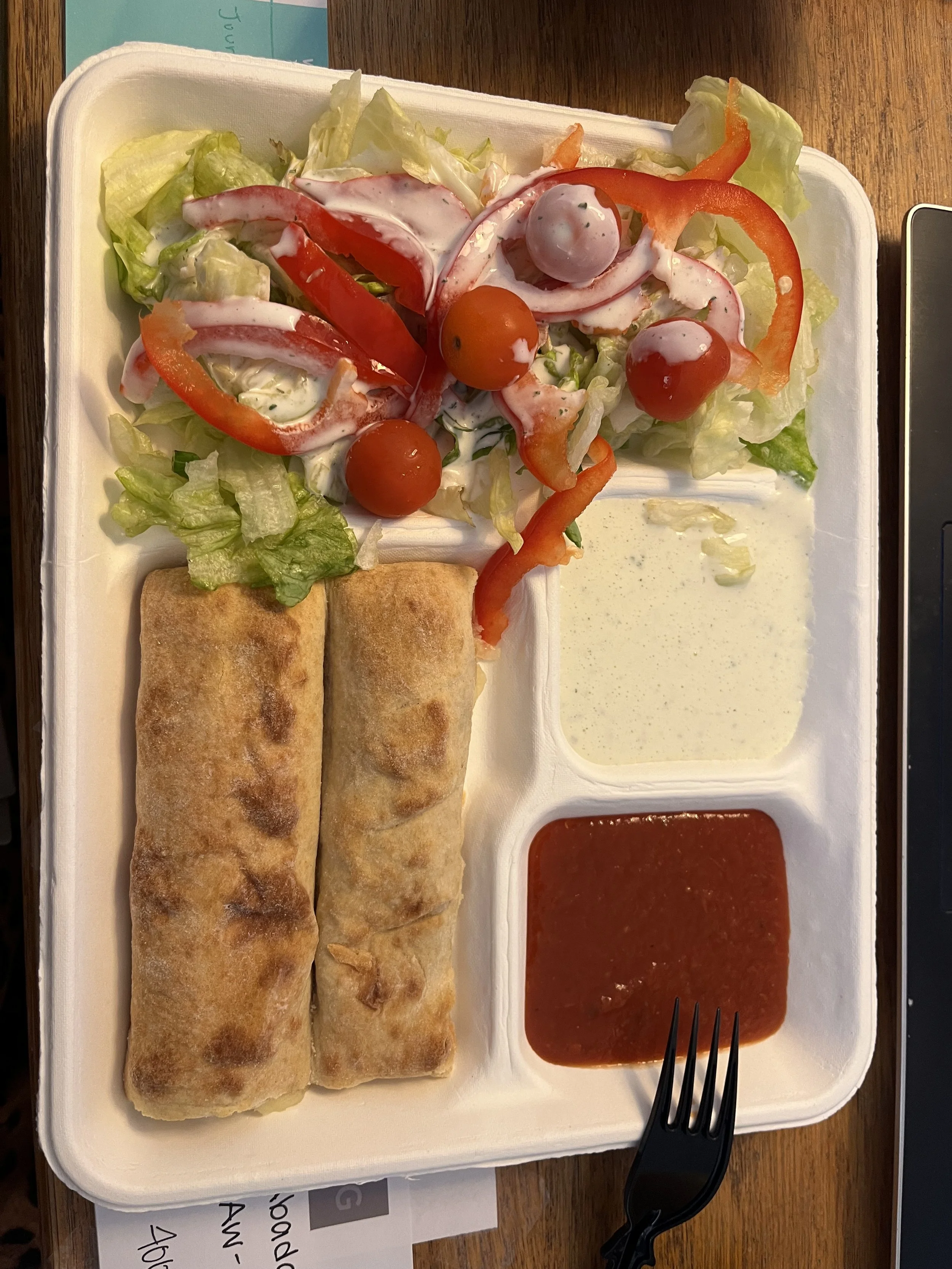 Cholula Mexican food container with two egg rolls, a side salad with cherry tomatoes, lettuce, red bell pepper, and dressing, and two types of dipping sauces.